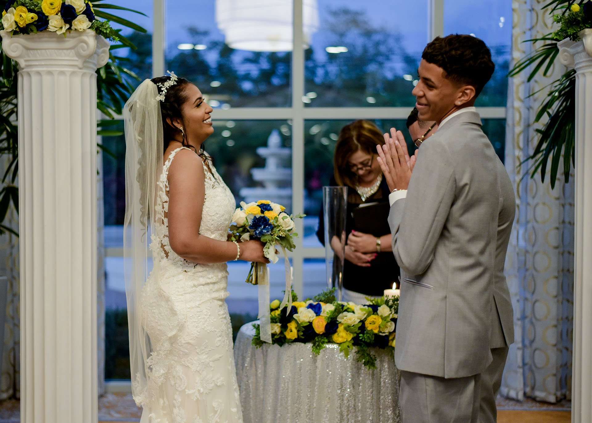 Bride in a lace gown laughs with her groom during an indoor ceremony with an officiant and blue and yellow bouquets