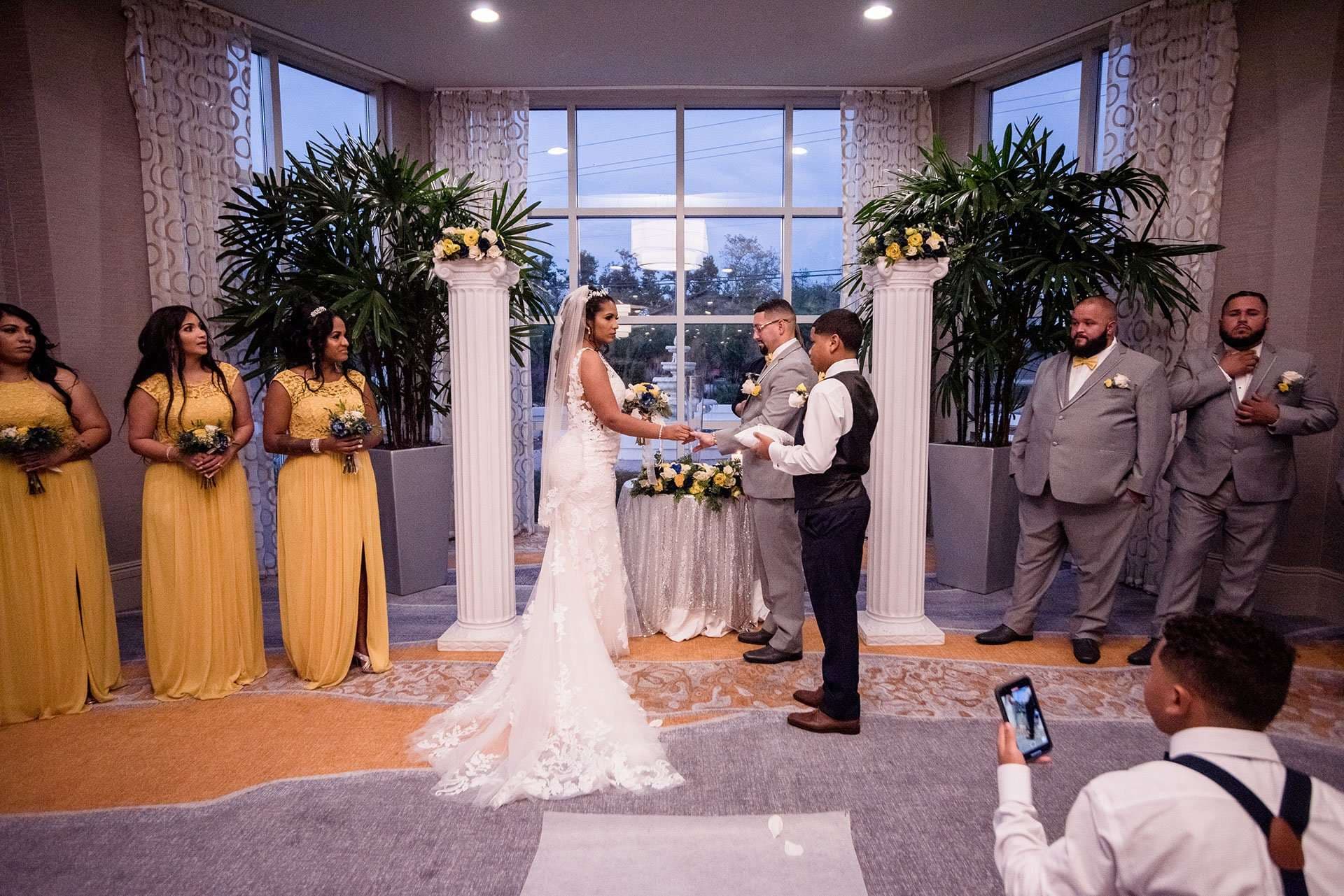 Bride and groom exchange vows before an officiant, flanked by bridesmaids in yellow gowns and groomsmen in gray suits