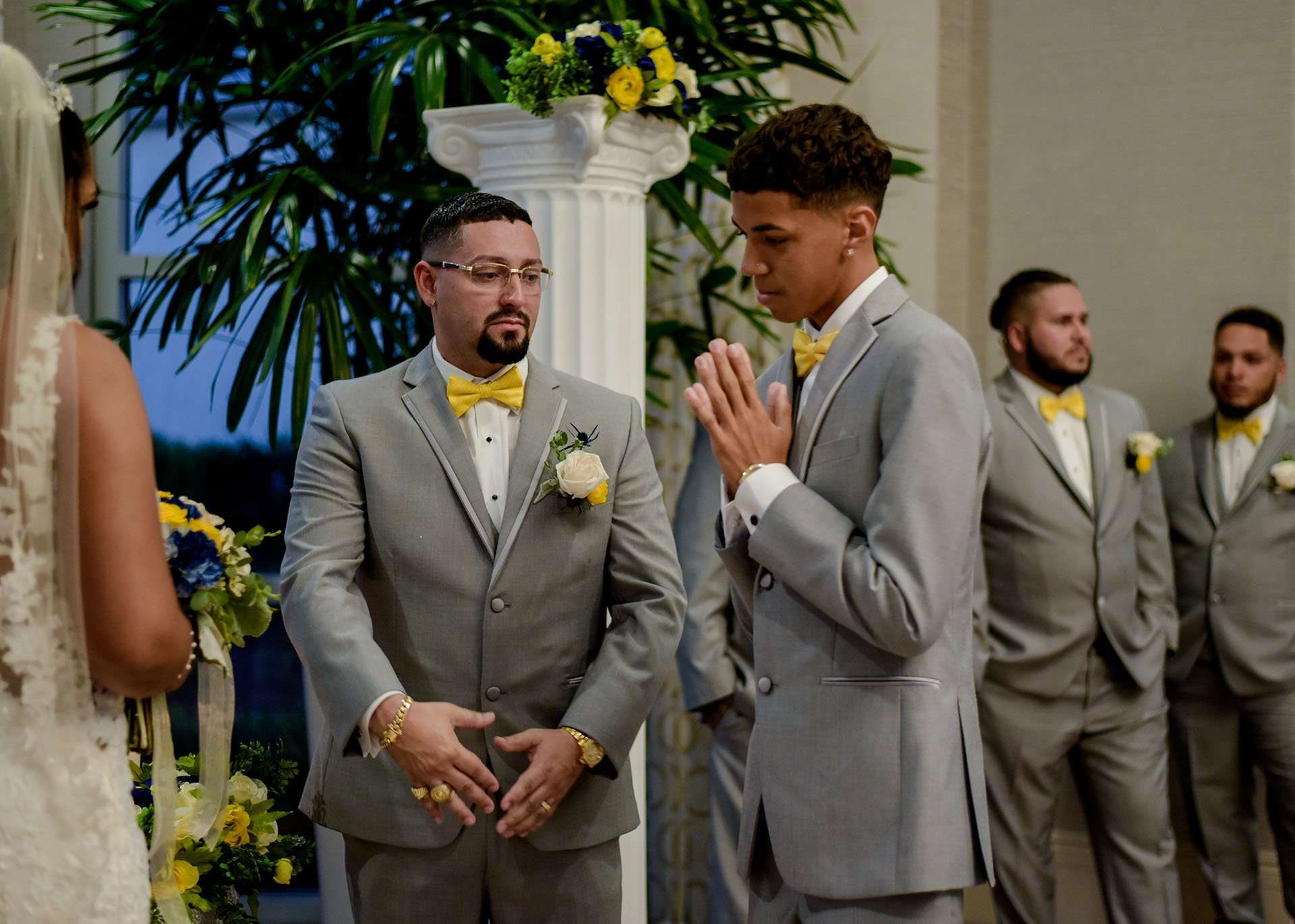 Groomsmen in gray suits and yellow bow ties stand at the altar as one holds his hands pressed together, facing the bride