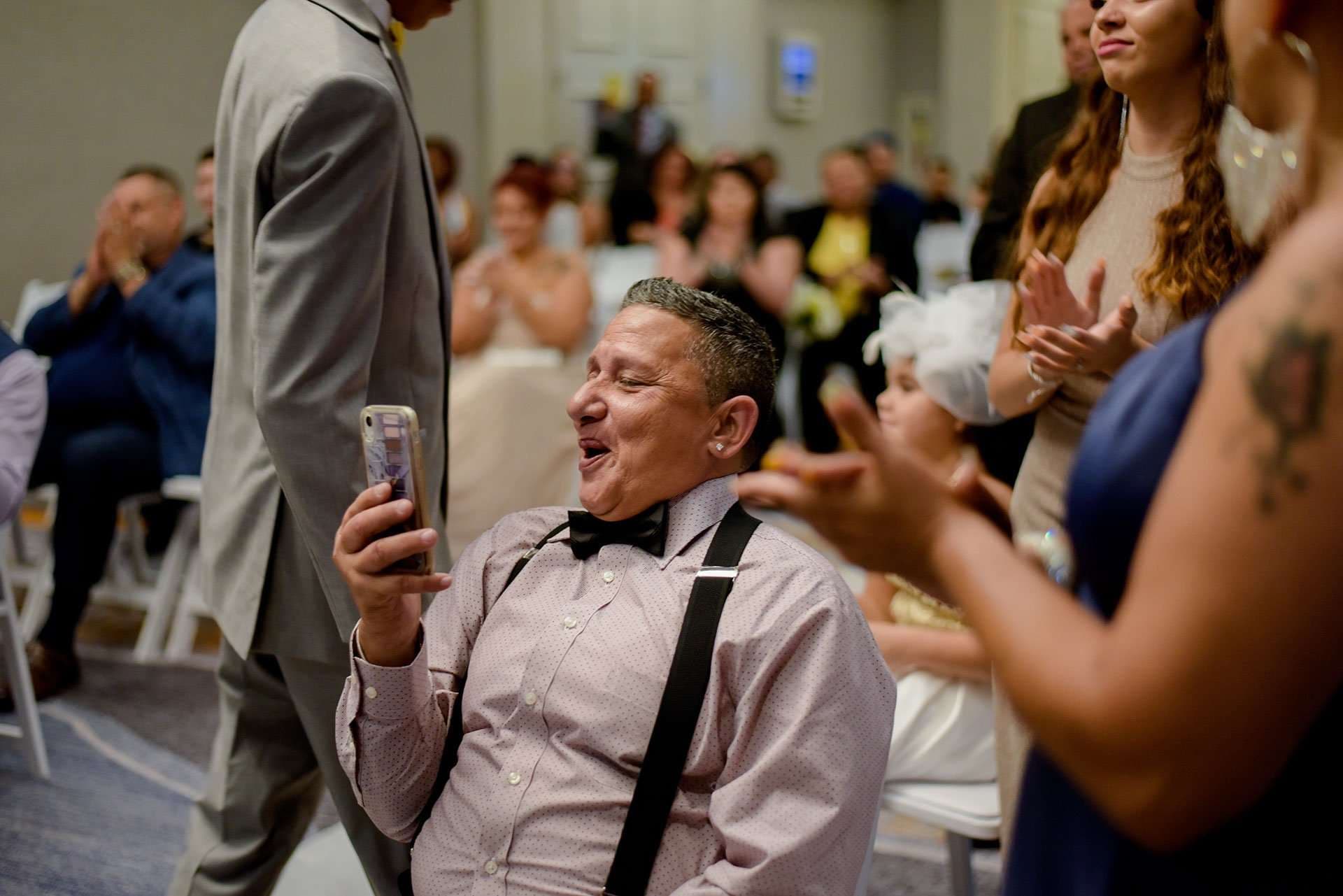 Smiling man in bow tie and suspenders holds up a phone while wedding guests around him applaud