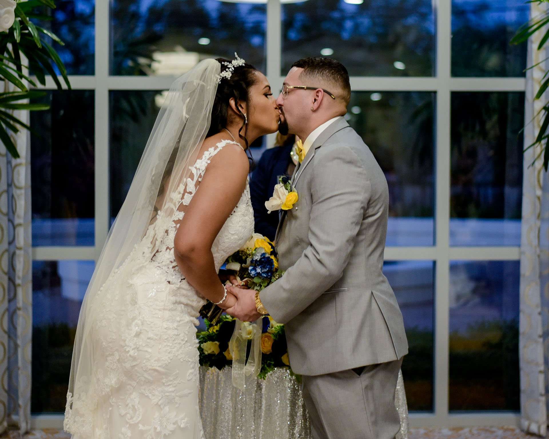 Bride in a lace gown and veil kissing her groom in a gray suit, holding a blue and yellow bouquet