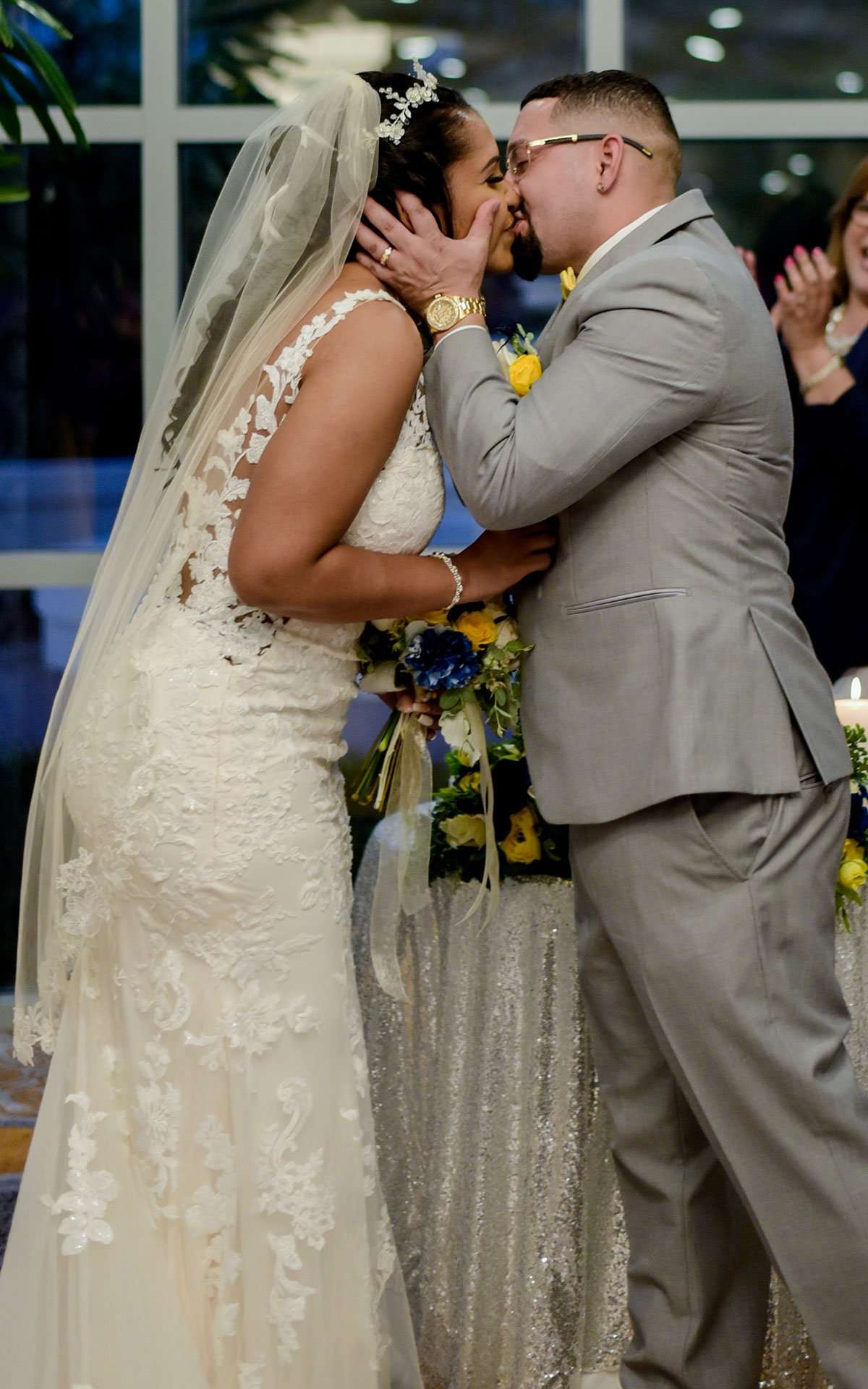 Bride in a lace gown and veil kissing her groom in a gray suit during the ceremony, guest applauding behind