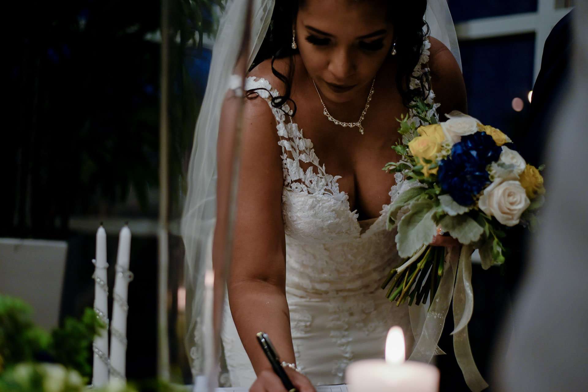 Bride in a white lace gown and veil signs a document, holding a blue and yellow bouquet beside lit candles