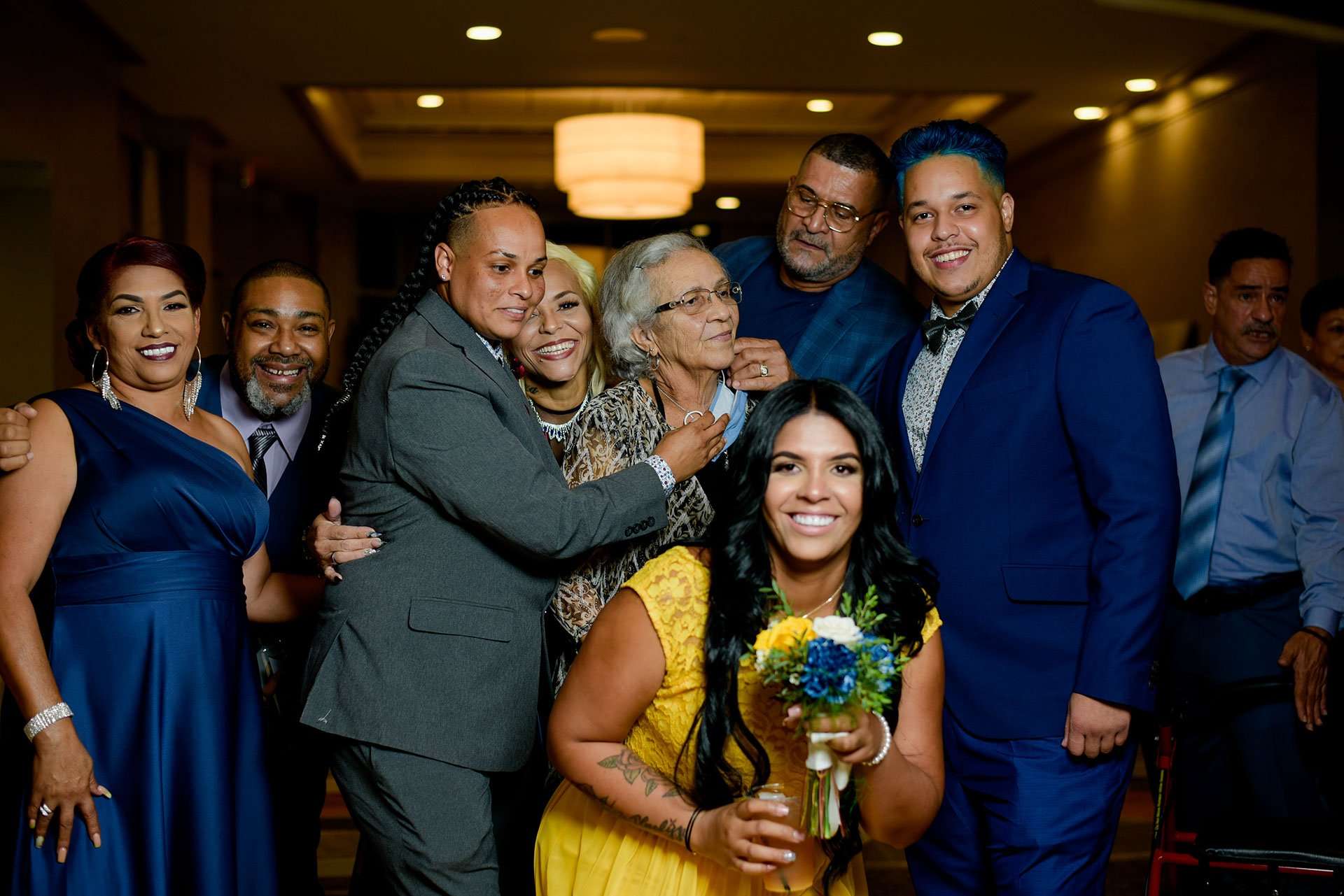 Smiling wedding guests in blue and gray formalwear gather around an older woman while a woman in yellow holds a bouquet