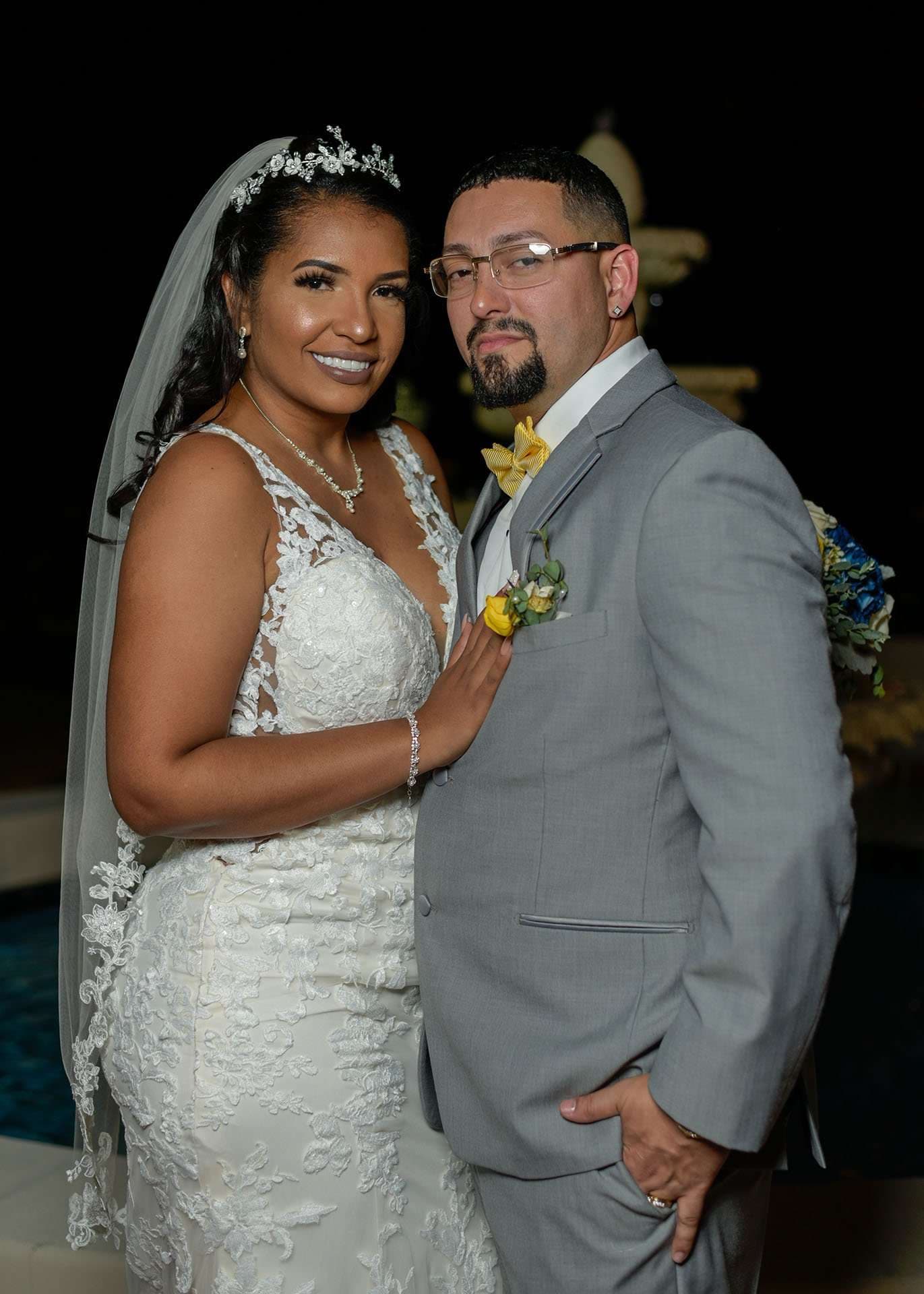 Bride in a sparkling tiara and lace gown posing with the groom in a gray suit at night