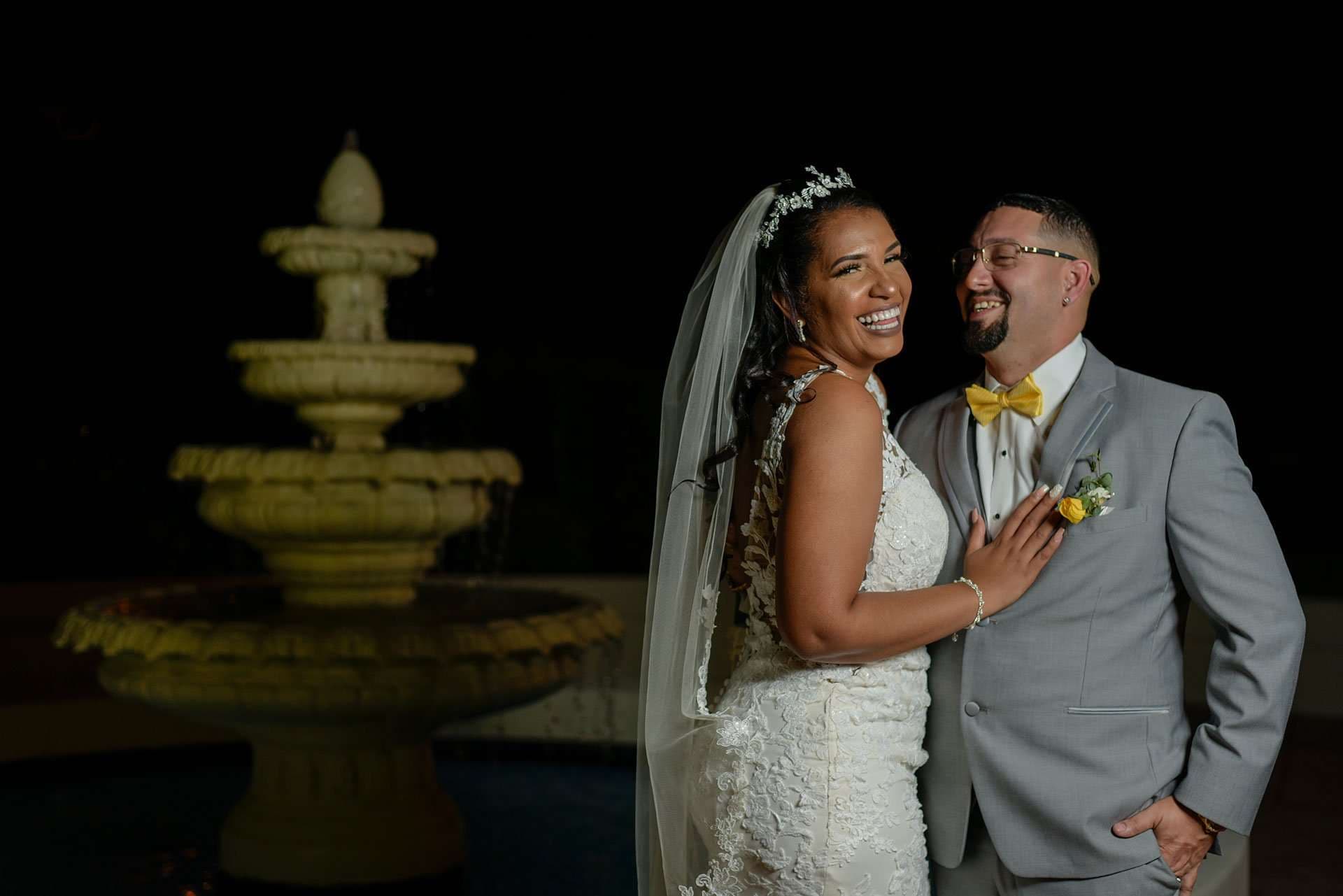 Laughing bride in a lace gown and veil embracing her groom in a gray suit and yellow bow tie by a lit fountain at night