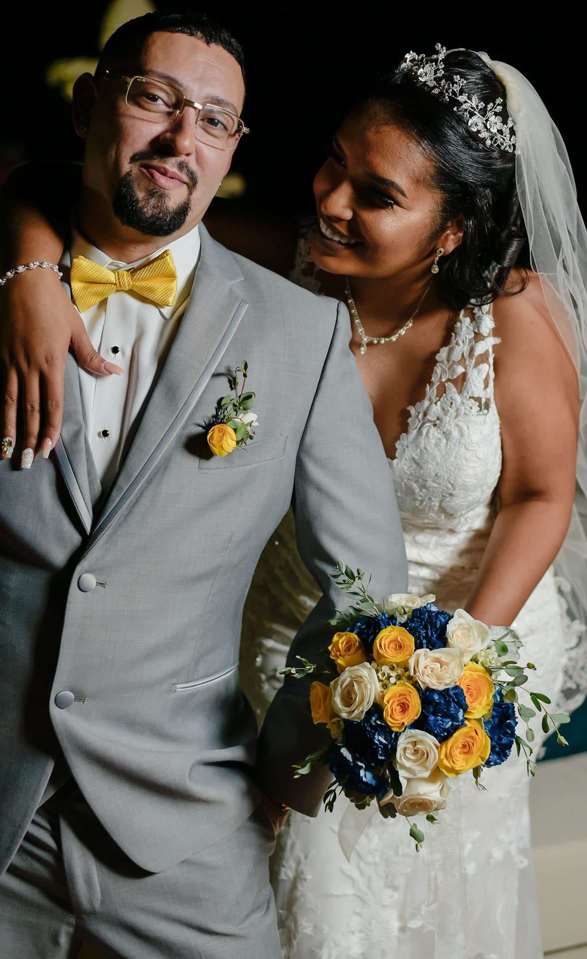 Smiling bride in a lace gown and veil leans on her groom in a gray suit and yellow bow tie, holding a bouquet