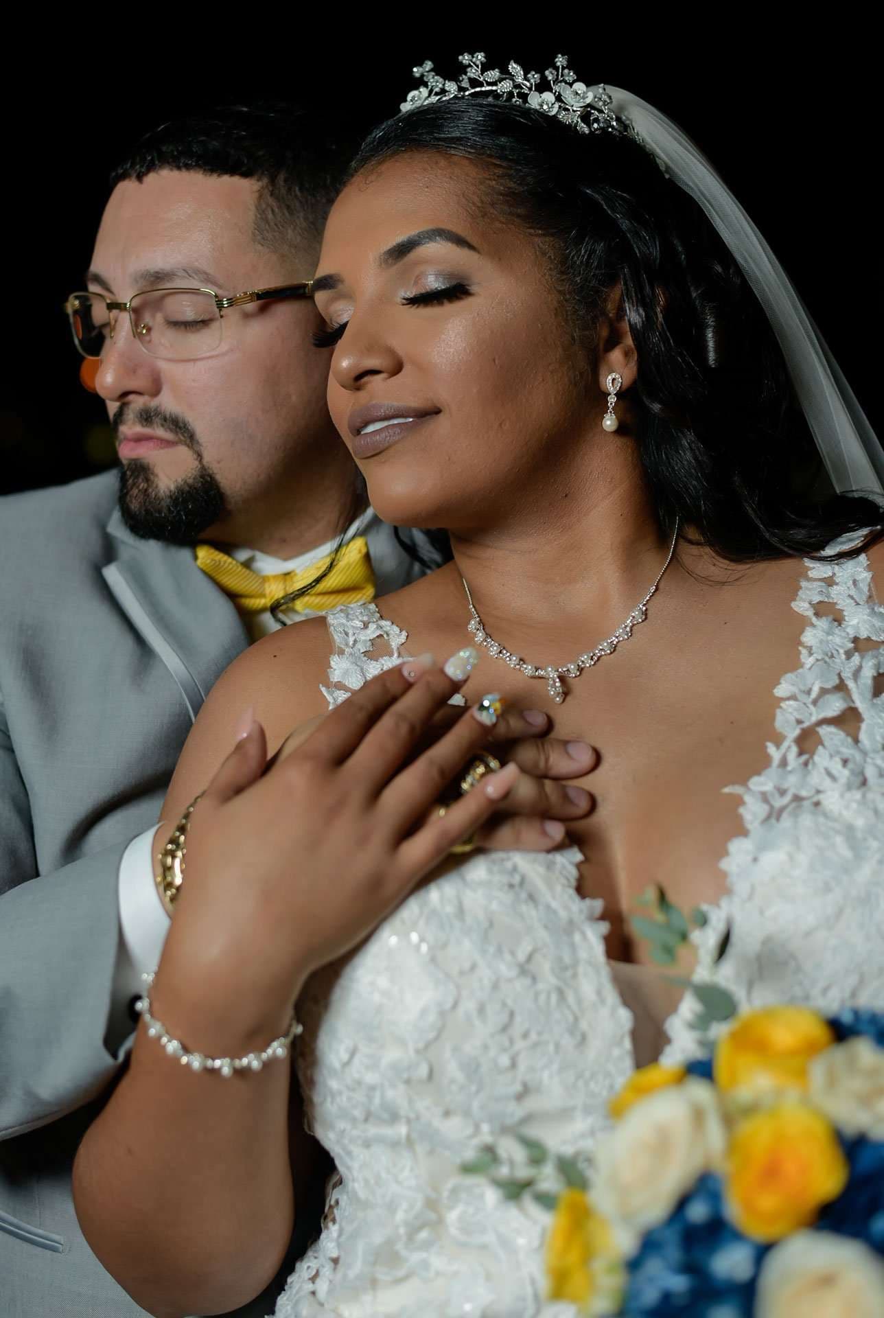 Bride in a lace gown and tiara with eyes closed as her groom in a yellow bow tie embraces her from behind