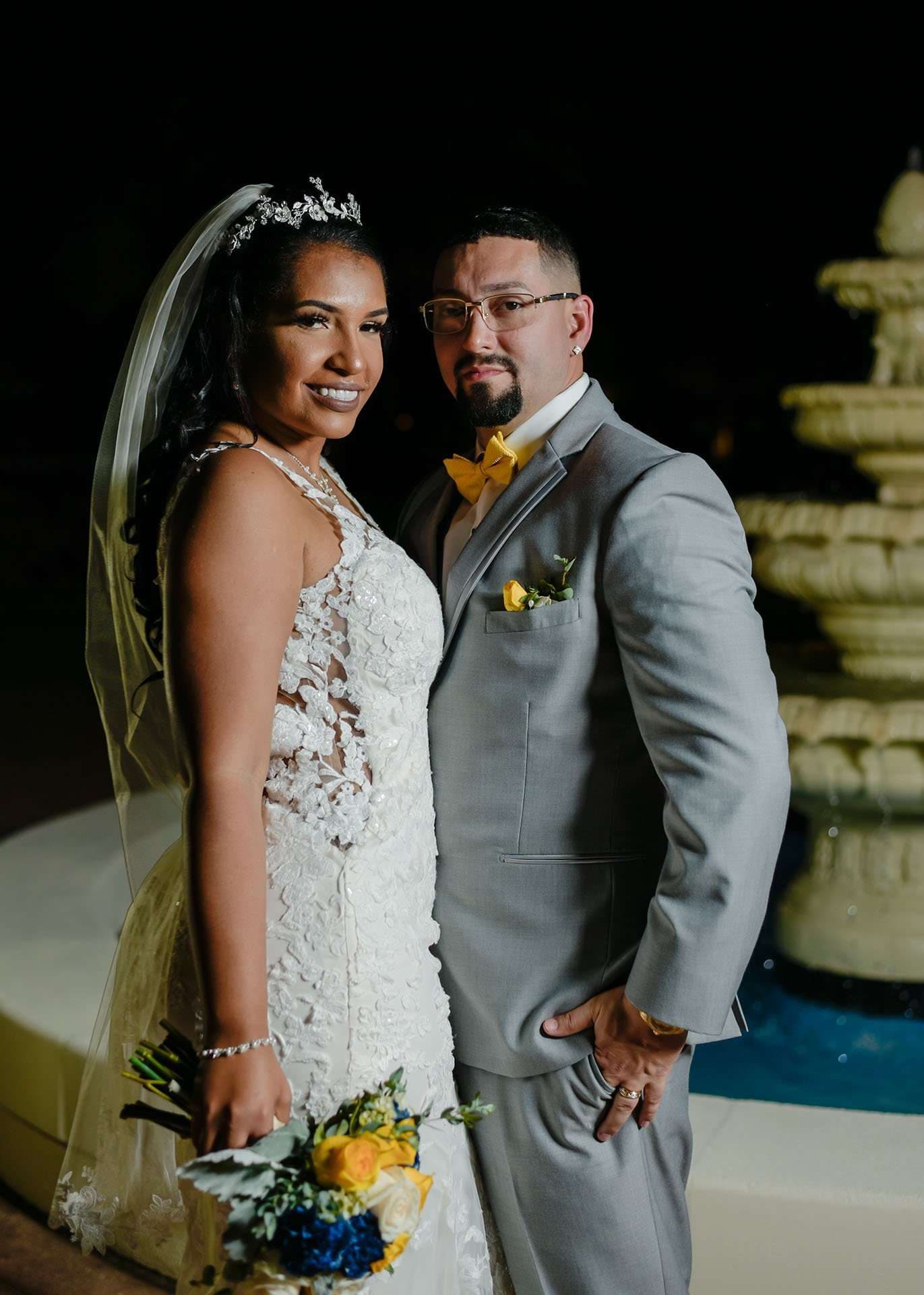 Bride in a white lace gown and veil beside a groom in a gray suit and yellow bow tie near a night fountain