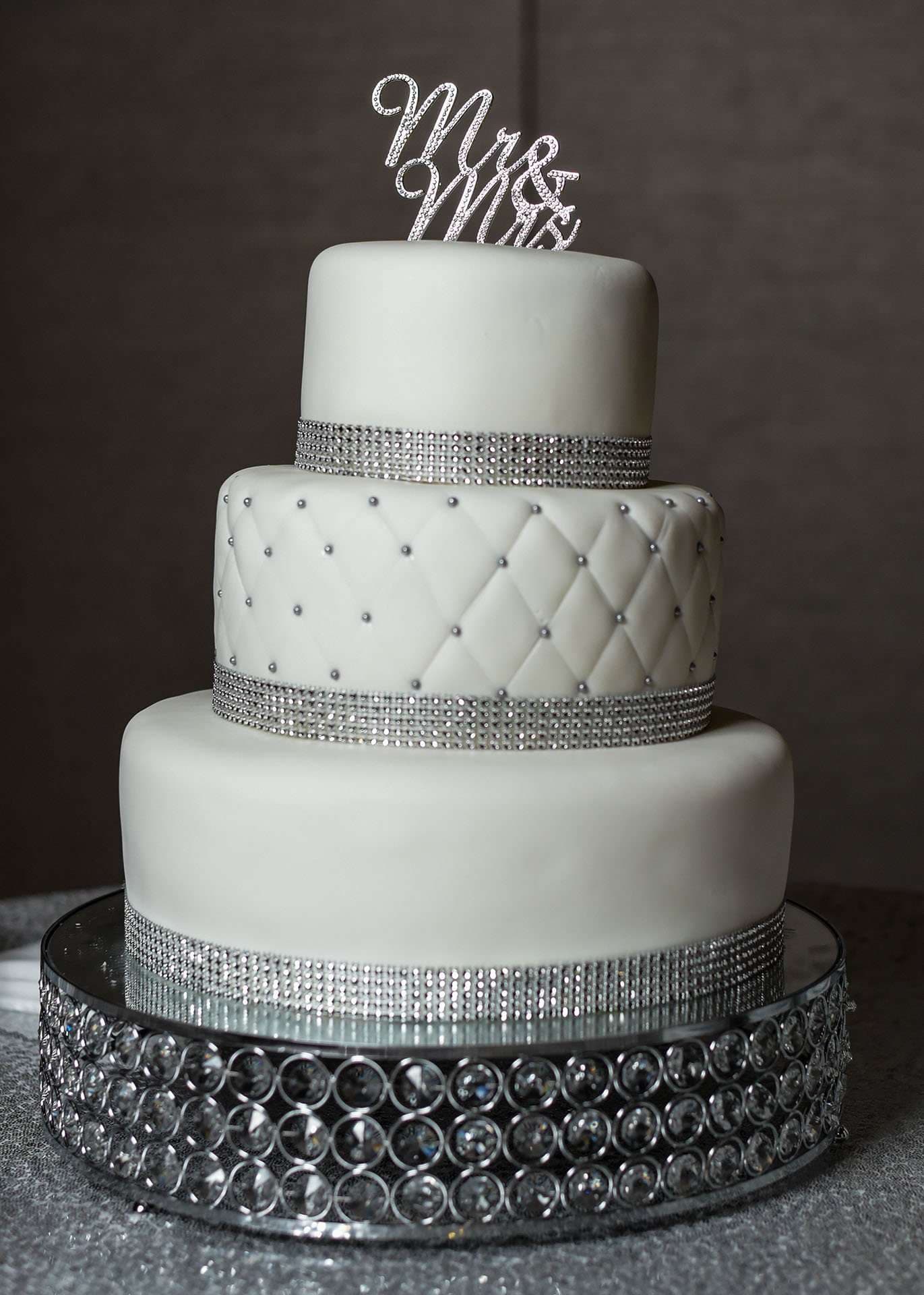 Three-tier white wedding cake with quilted middle tier, rhinestone bands and Mr & Mrs topper on a crystal stand