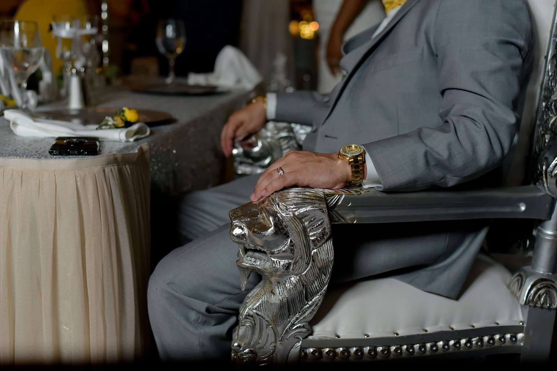 Man in a gray suit and gold watch seated on an ornate silver throne chair with a lion armrest at a reception table
