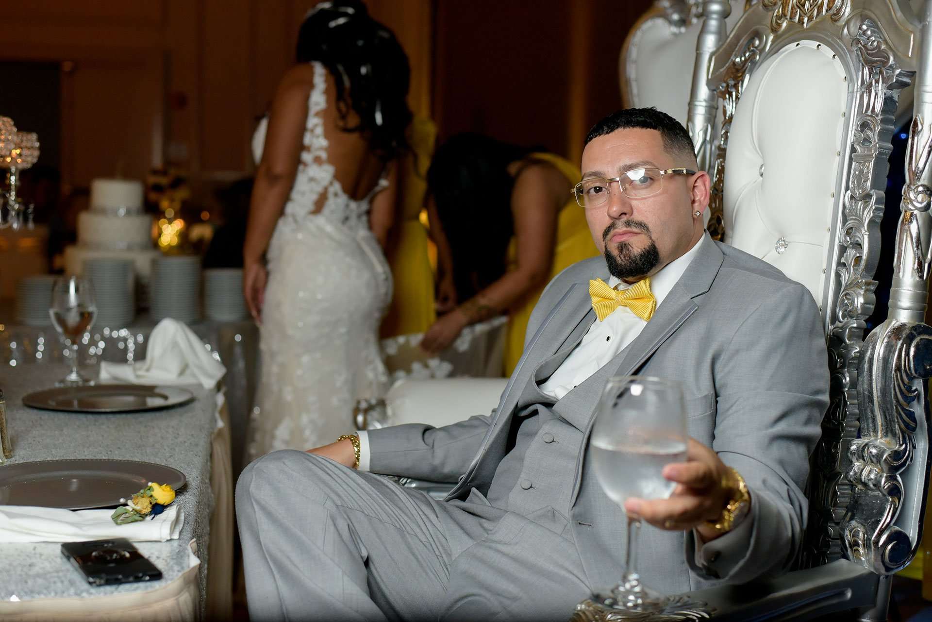 Man in gray suit and yellow bow tie holding a wine glass on an ornate silver throne chair at a reception