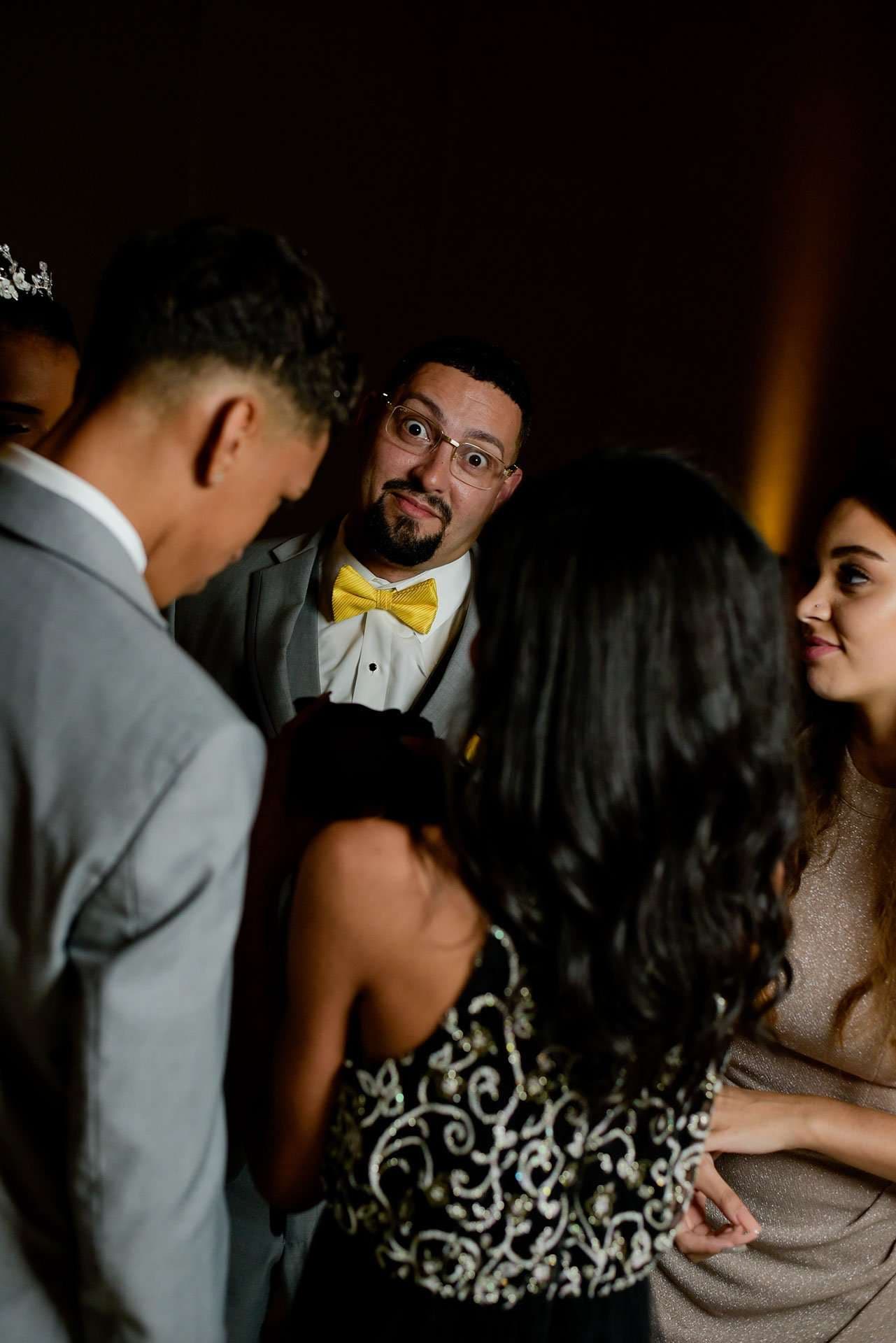 Man in glasses and yellow bow tie makes a funny wide-eyed face behind guests at a dimly lit reception