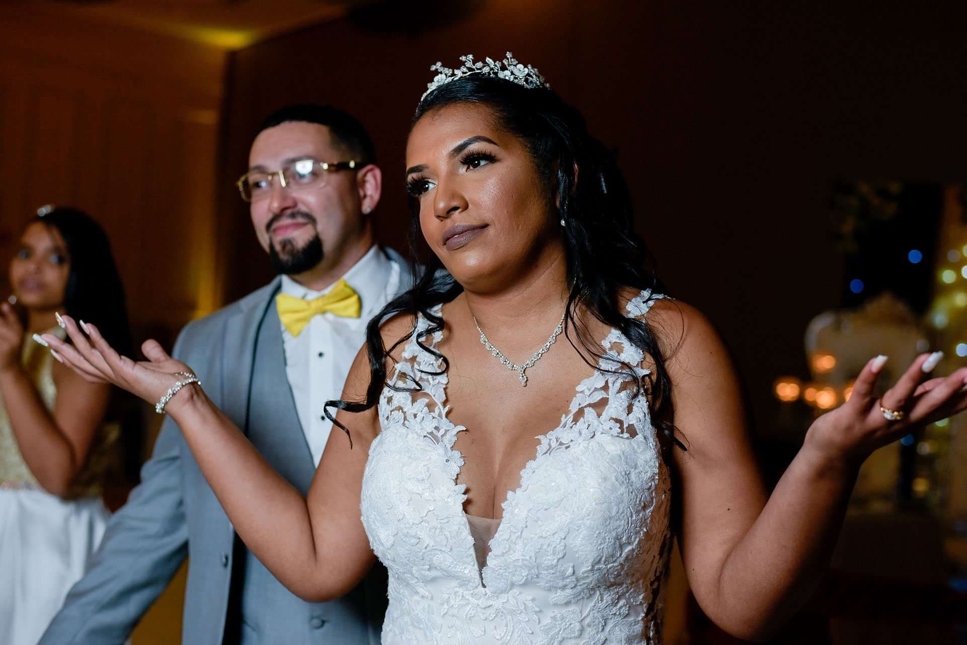 Bride in a white lace gown and tiara shrugs with open hands while a groom in a gray suit and yellow bow tie stands behind