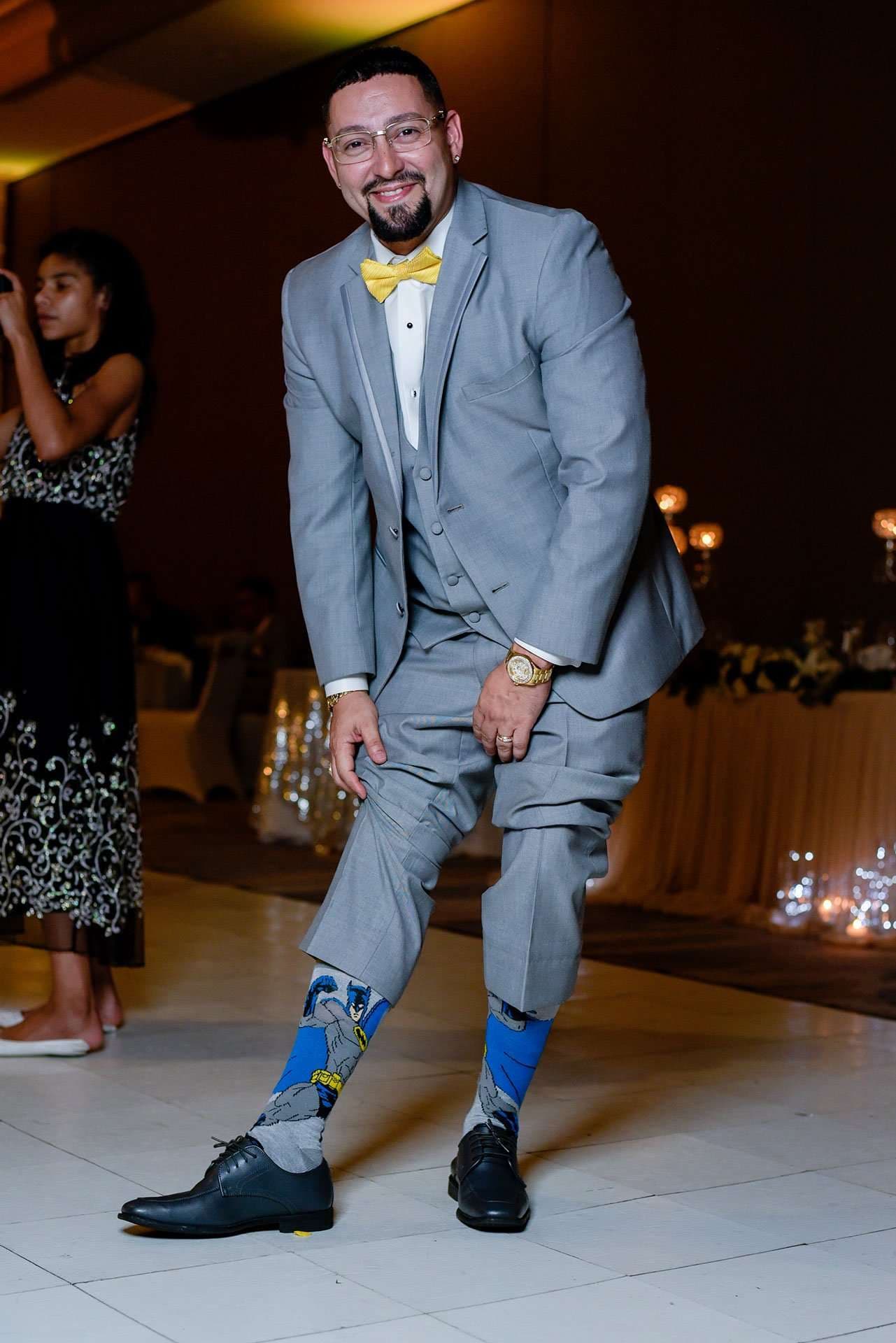 Smiling man in gray suit and yellow bow tie lifts his pant legs to show Batman socks on a reception dance floor