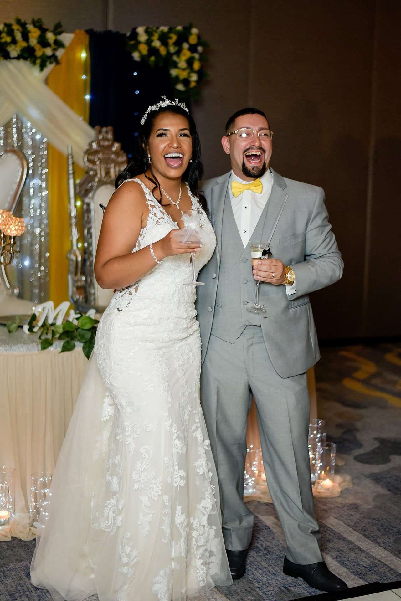 Laughing couple in a white lace gown and gray suit with yellow bow tie holding champagne glasses at their reception