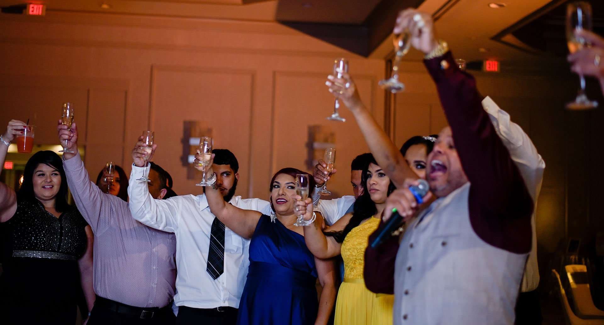 Wedding guests raise champagne glasses in a toast while a man in a gray vest sings into a microphone
