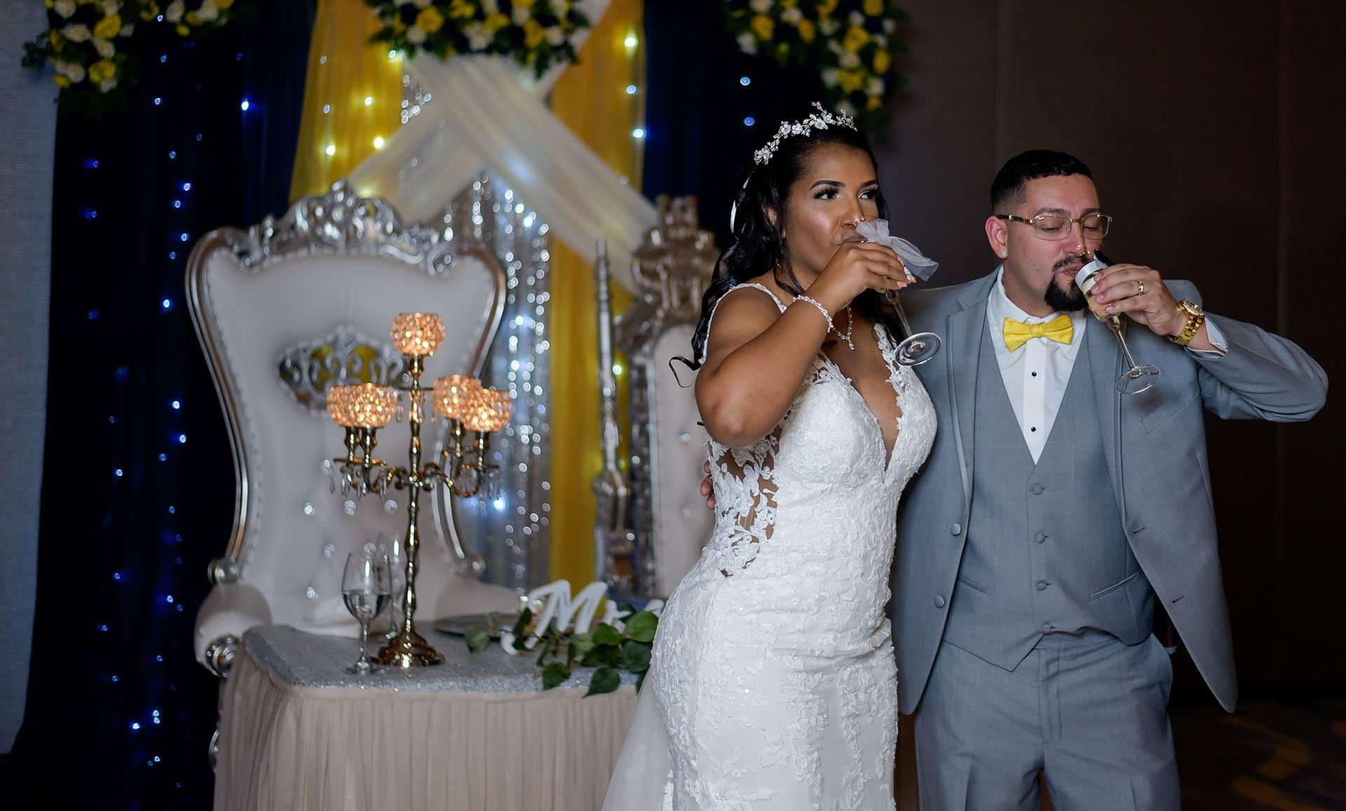 Bride in a white lace gown and groom in a gray suit with yellow bow tie toasting beside an ornate white throne