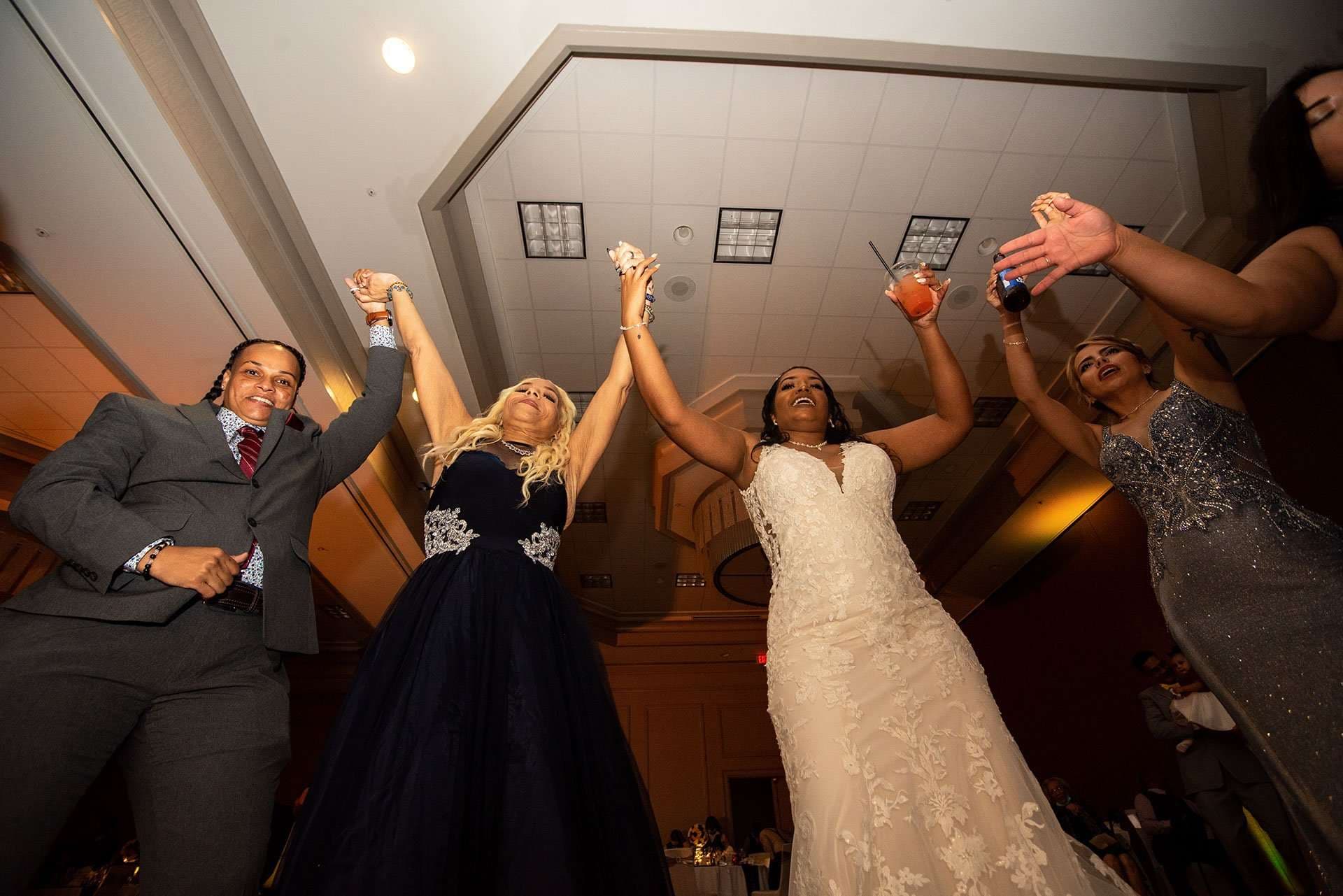 Bride in white lace gown and guests raising joined hands and drinks while dancing at a reception, shot from below