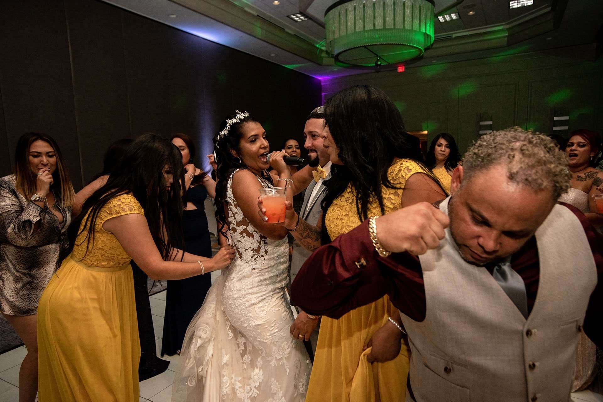 Bride in a lace gown sings into a microphone while bridesmaids in yellow dresses dance around her at the reception