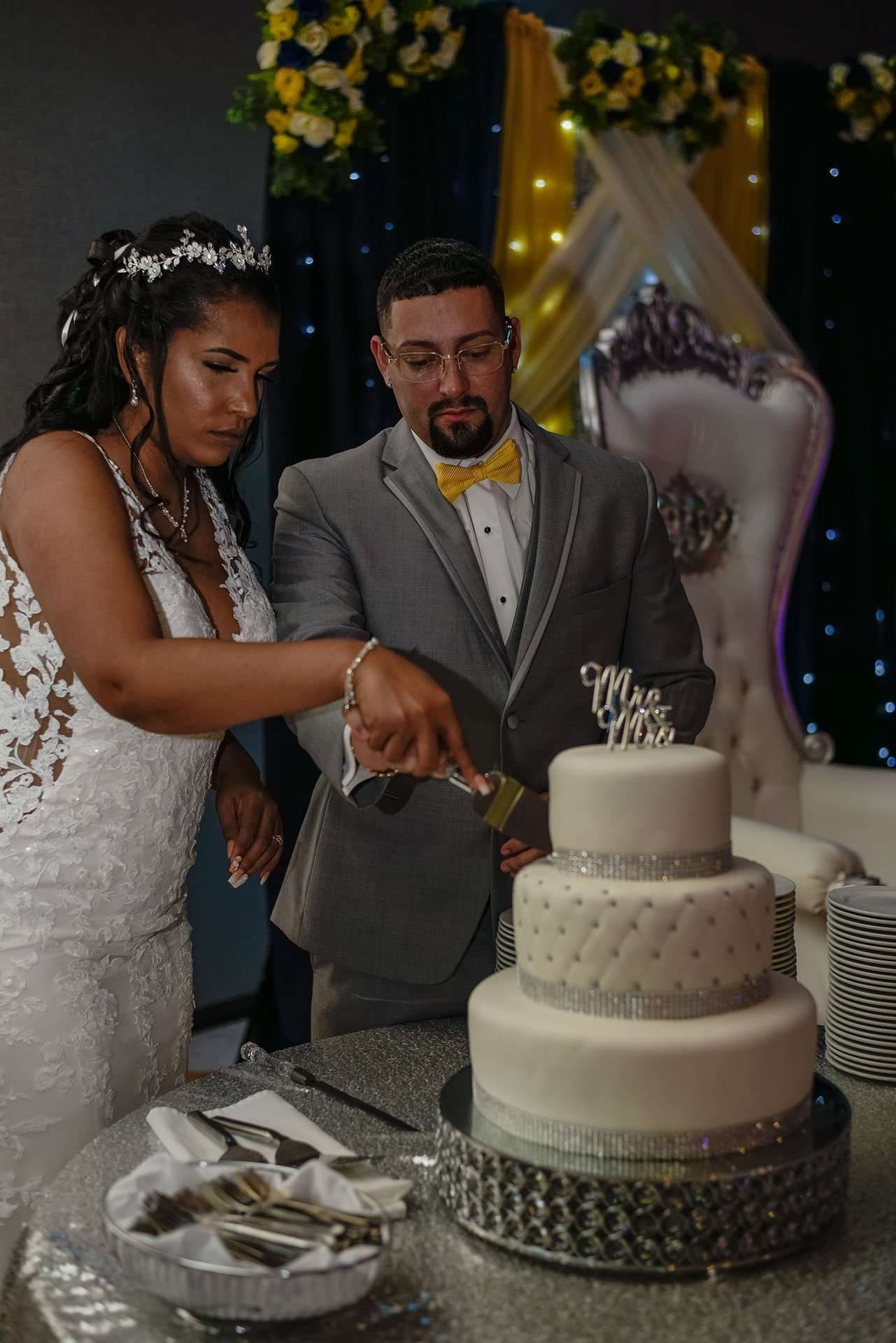 Couple cutting a three-tier white wedding cake topped with a Mr & Mrs sign, bride in lace gown, groom in gray suit