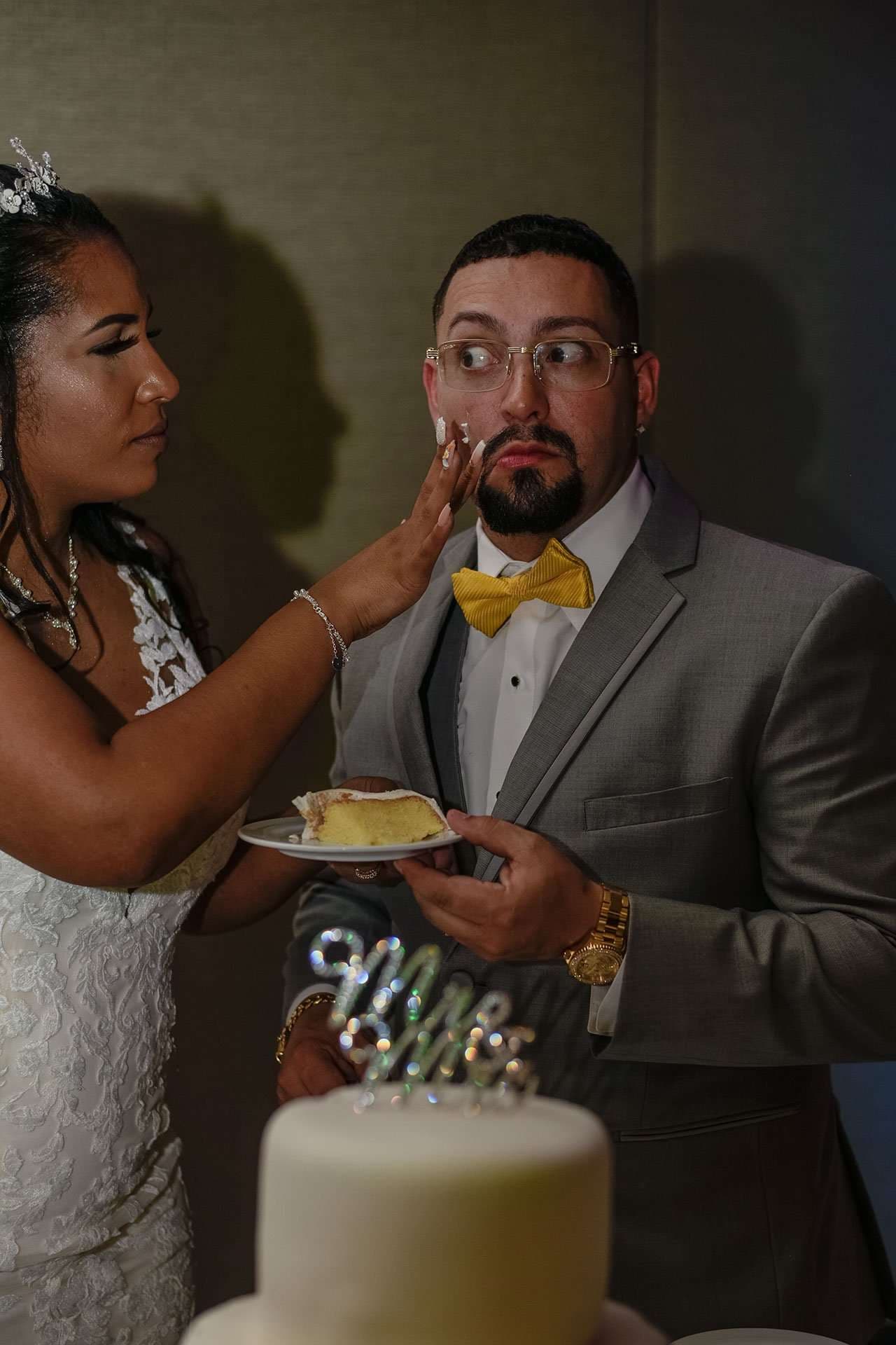 Bride in white lace gown playfully touching her bearded groom's face as he holds a cake slice, looking wide-eyed