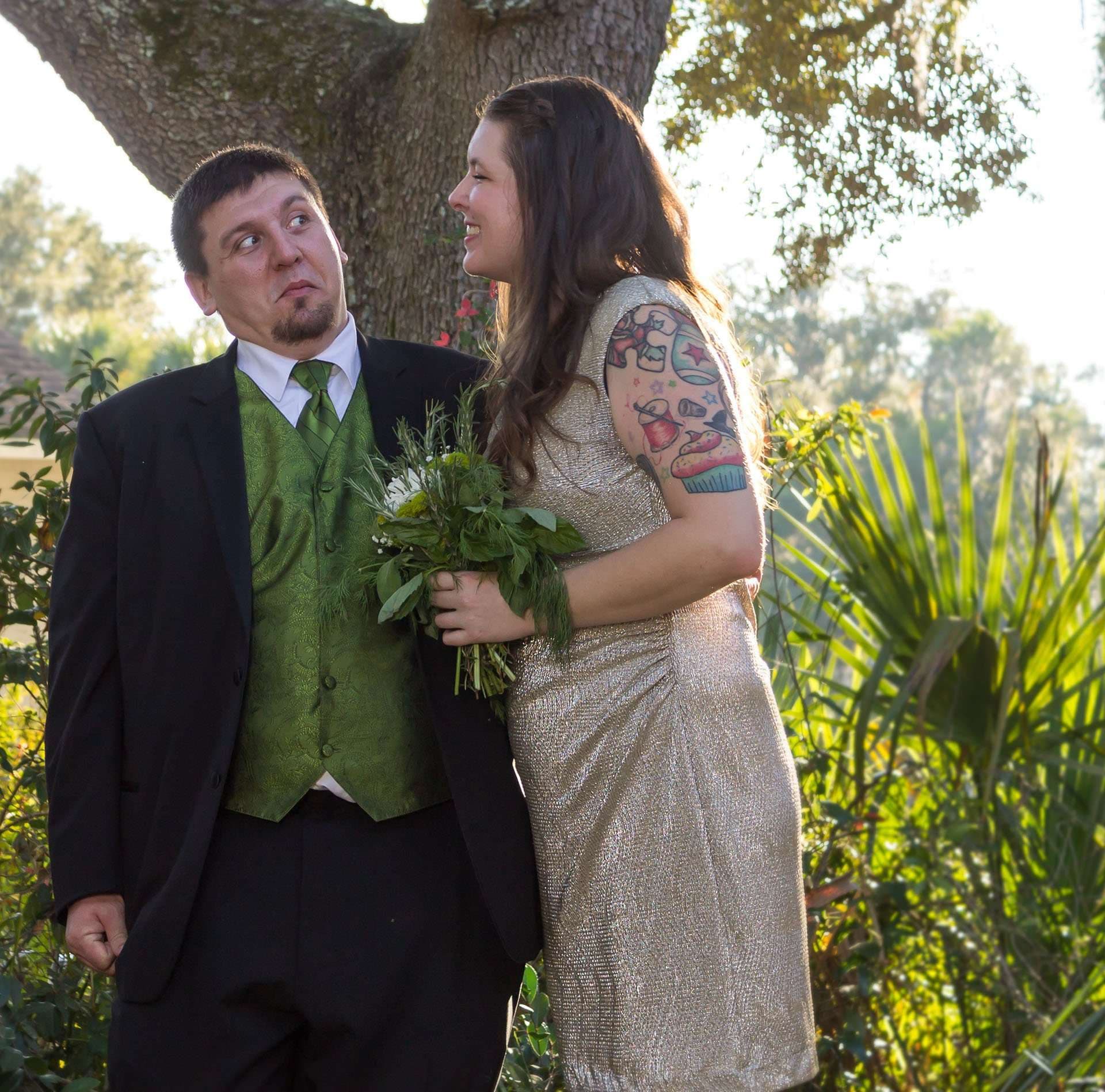 Smiling tattooed bride in a champagne dress holding a bouquet while the groom in a green vest glances sideways