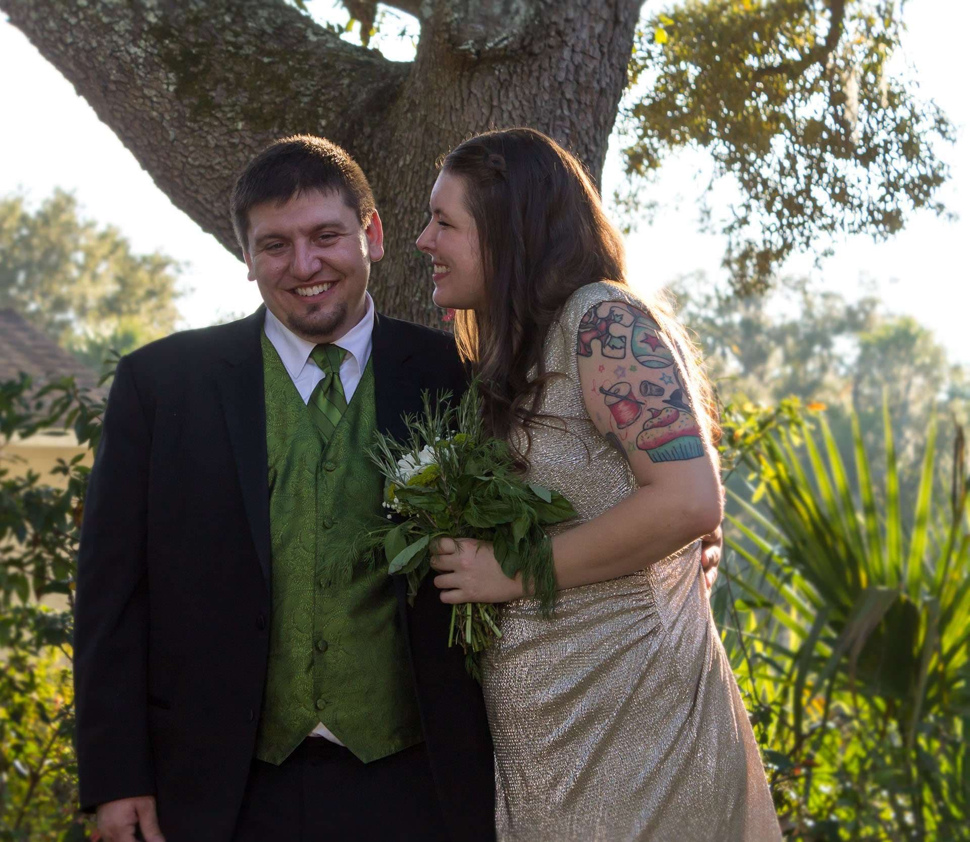 Smiling couple beside an oak tree, man in green vest and tie, tattooed woman in a gold dress holding greenery bouquet