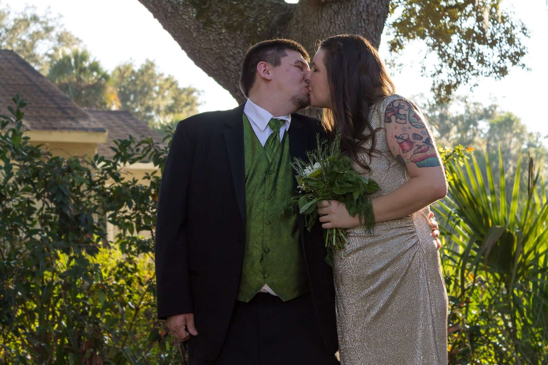 Couple kissing outdoors under an oak tree, he in a black suit and green vest, she in a sparkly gown holding greenery