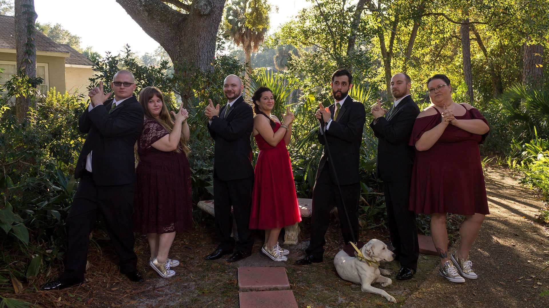 Wedding party in dark suits and burgundy and red dresses striking playful finger-gun poses in a garden, dog lying nearby