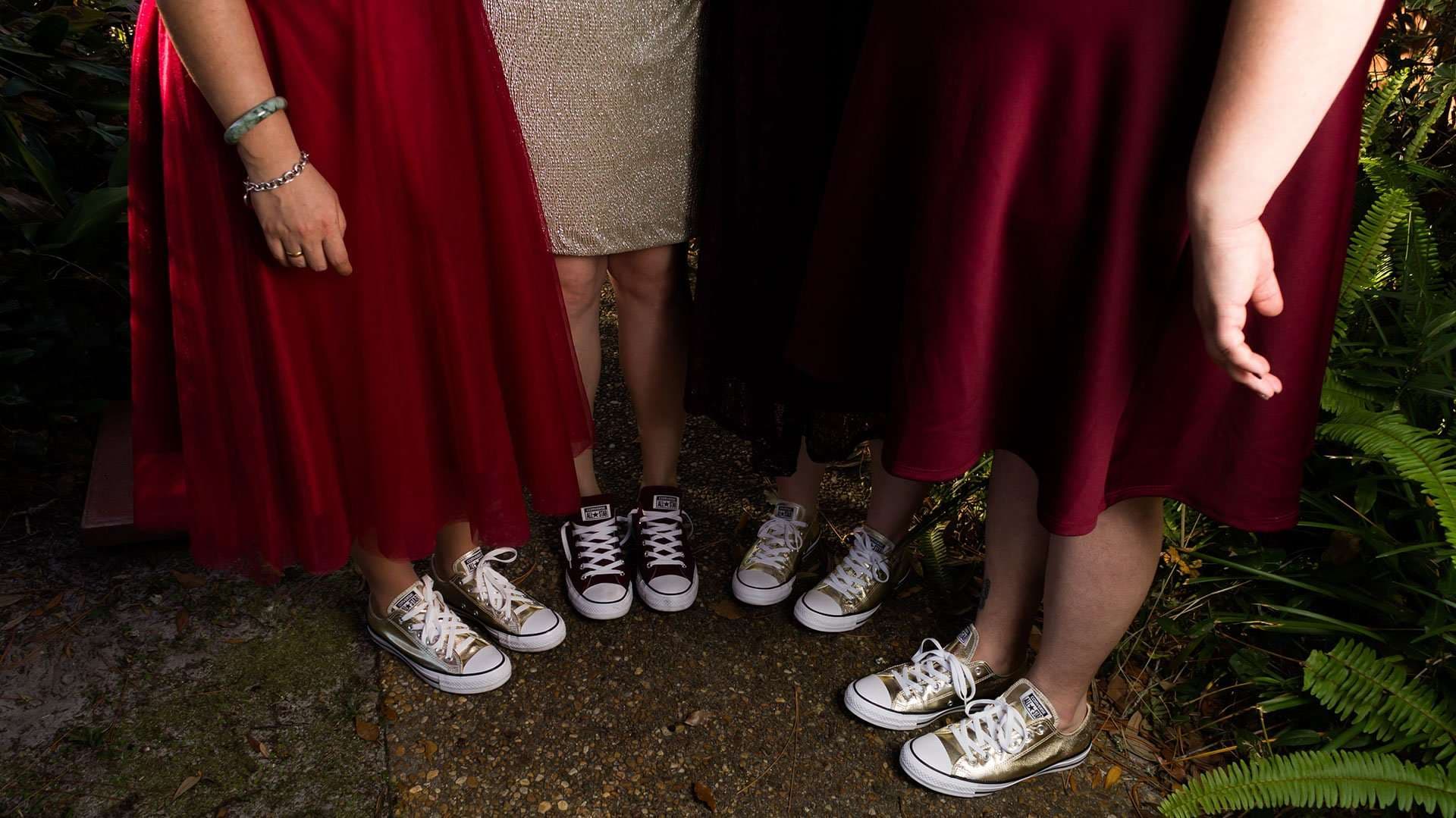 Four women in red and gold dresses standing together, each wearing matching Converse sneakers, beside ferns