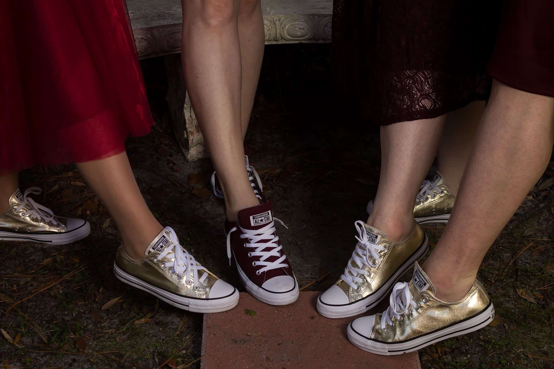 Four people's legs and low-top sneakers, three pairs metallic gold and one maroon, posed together on the ground outdoors