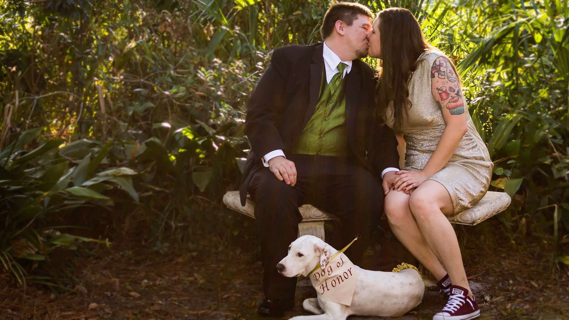 Couple kissing on a garden bench while their dog rests at their feet wearing a Dog of Honor sign