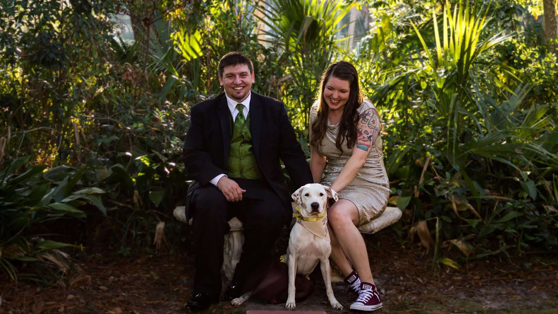 Smiling couple seated on a stone bench in a leafy garden with a white dog wearing a yellow bandana between them