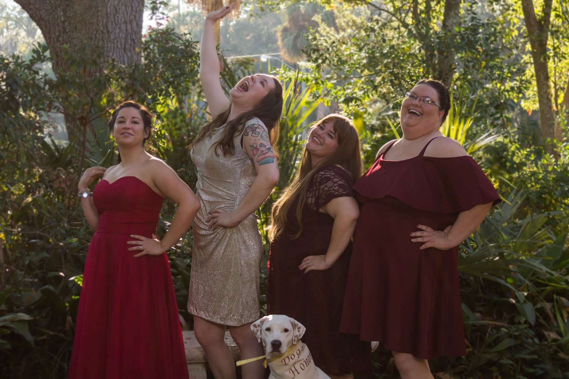 Four bridesmaids in red and gold dresses laughing and posing in a garden beside a dog wearing a Dog of Honor sign