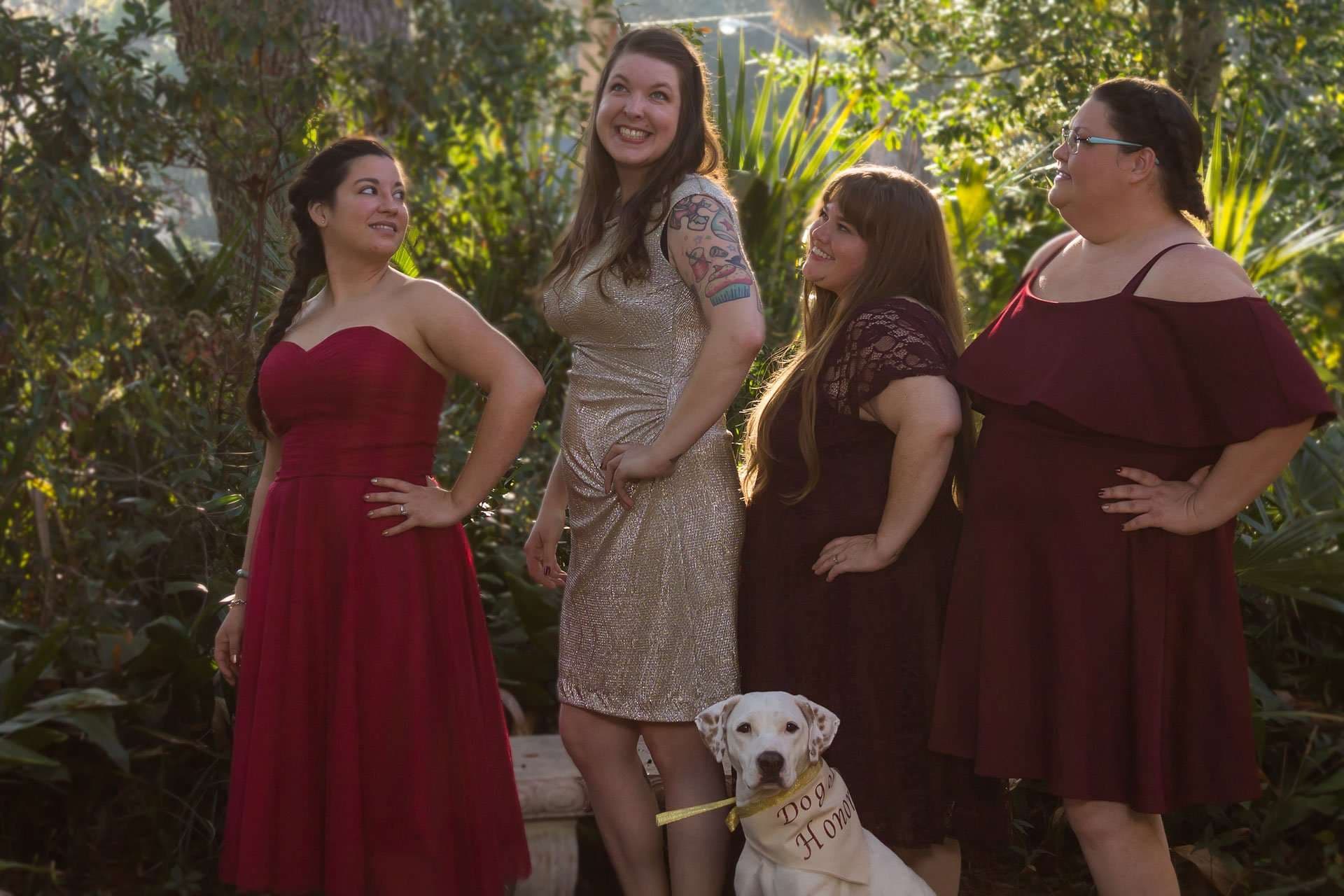 Four women in red and gold dresses pose in a sunlit garden beside a white dog wearing a Dog of Honor sign