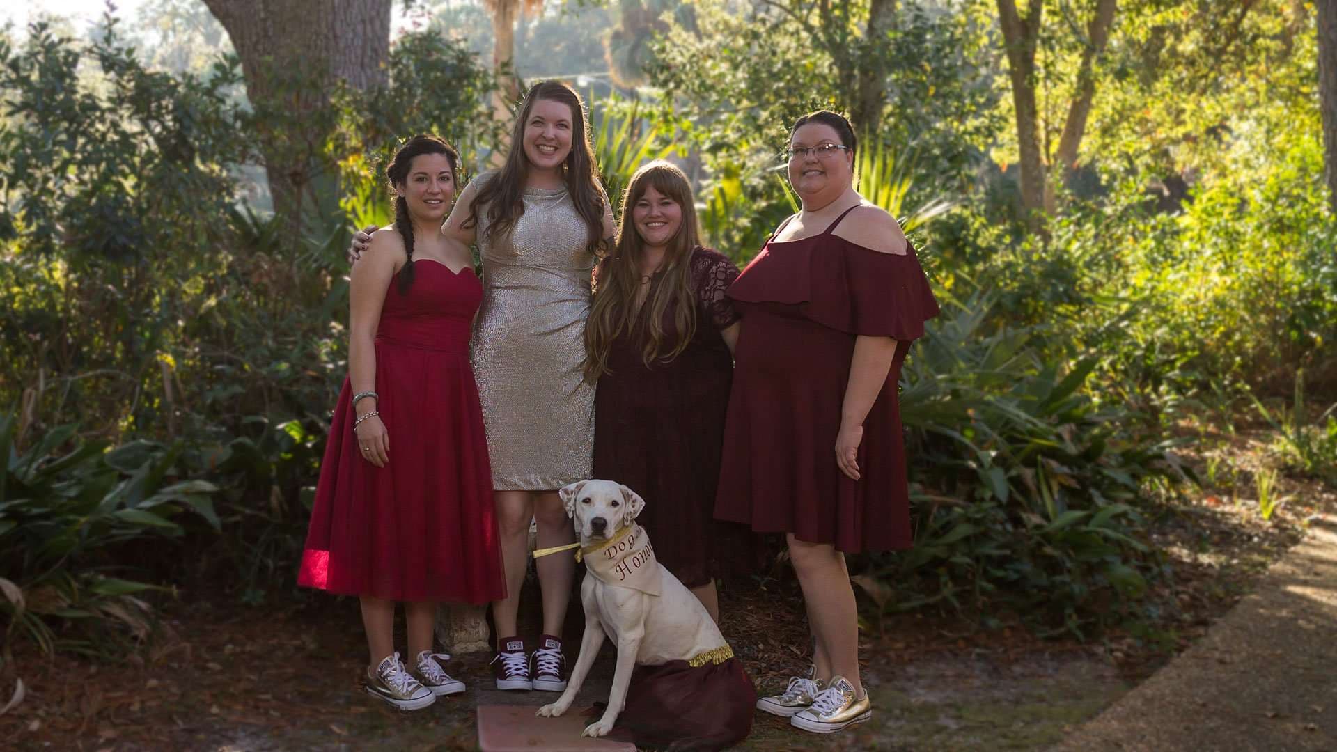 Four women in burgundy and champagne dresses and sneakers pose in a garden with a white dog wearing a sash