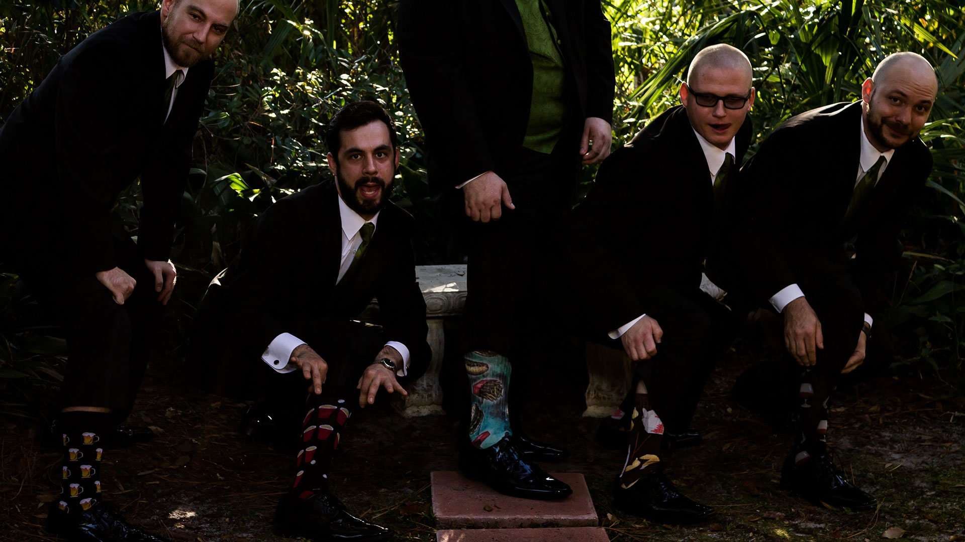 Four groomsmen in dark suits posing around a stone bench outdoors, showing off colorful patterned socks