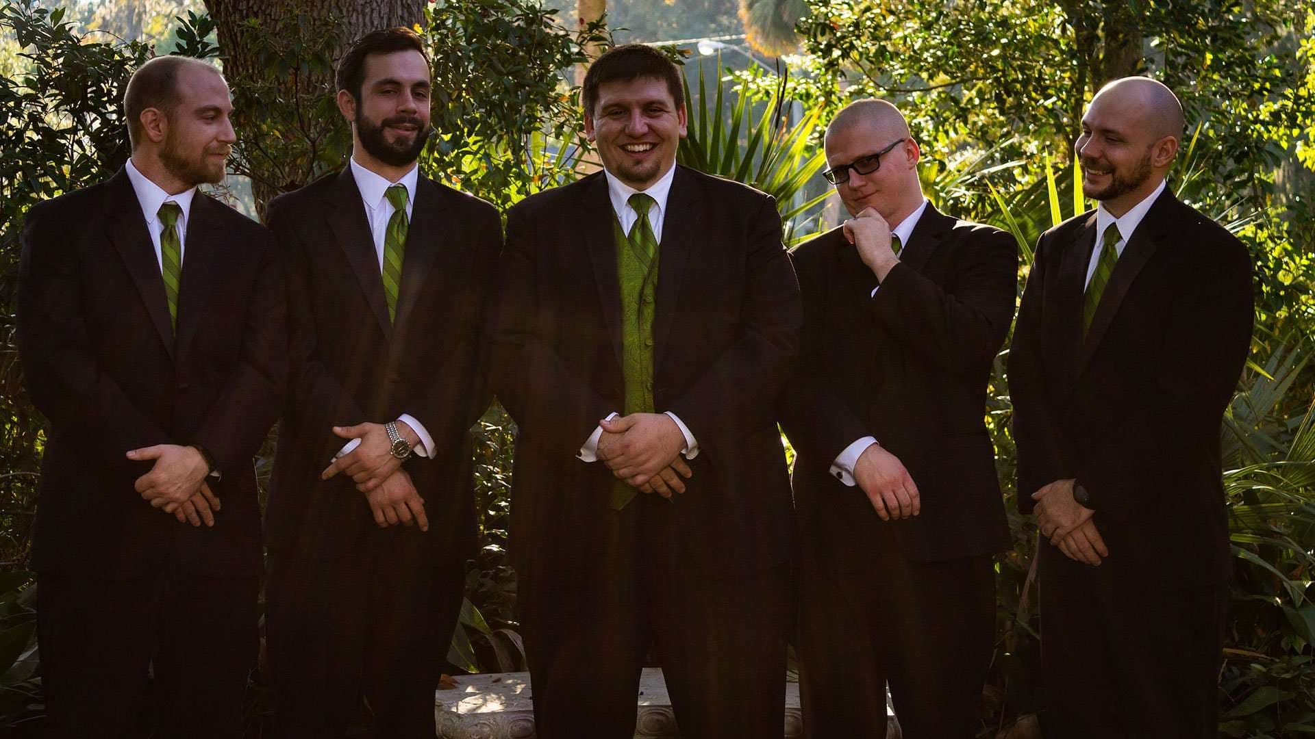 Five groomsmen in dark suits and green ties standing in a row in a leafy garden, the center one smiling