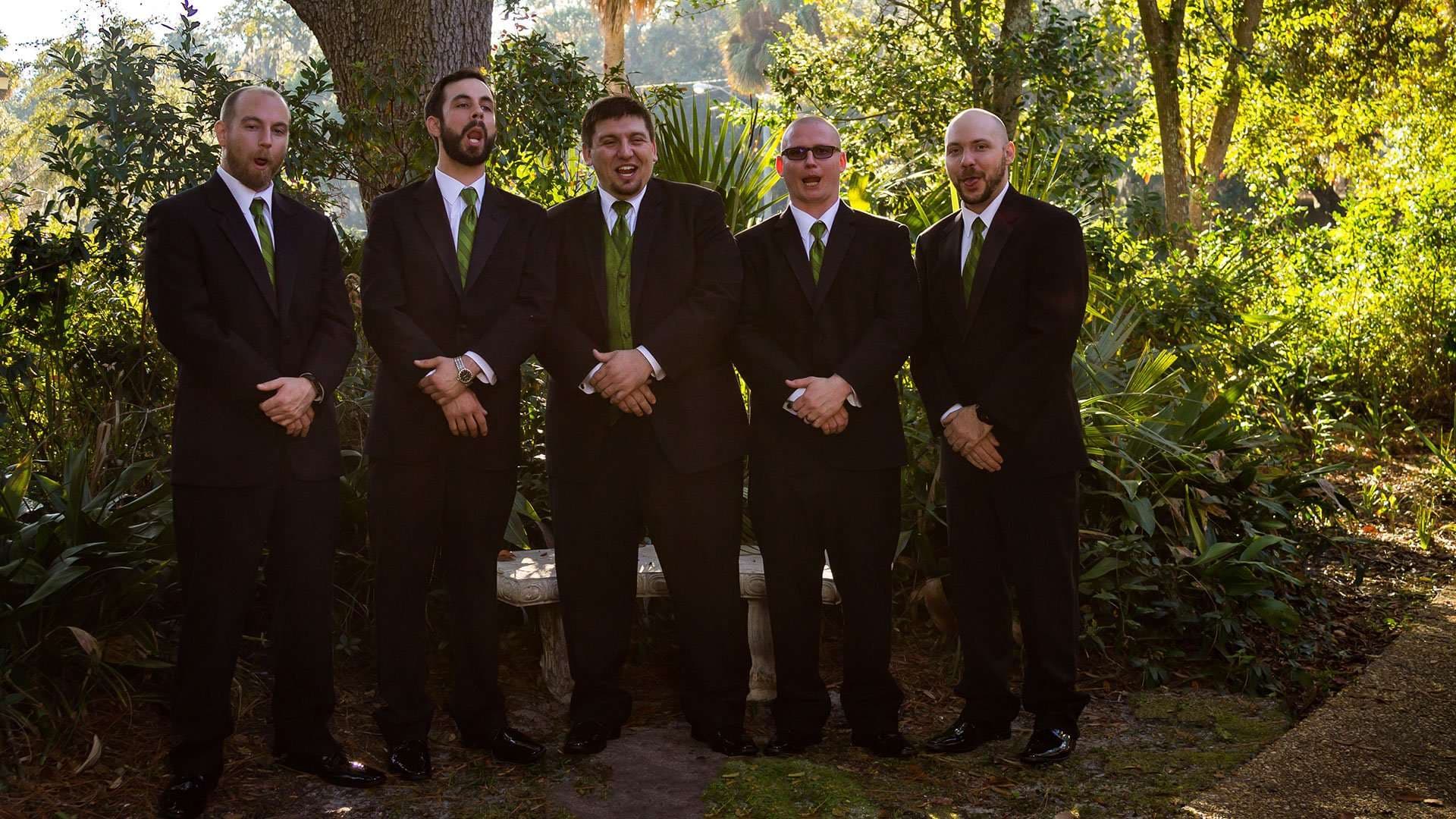 Five men in dark suits and green ties standing side by side in a sunlit garden, hands clasped in front