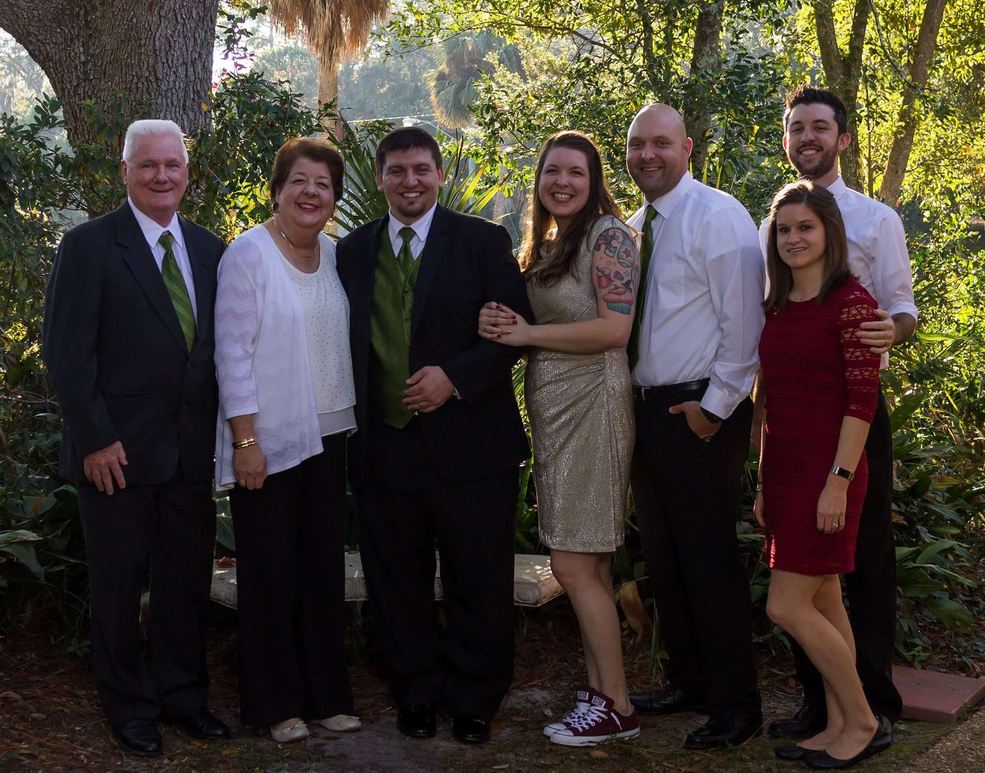 Six family members and friends in dressy attire pose close together outdoors among trees and greenery