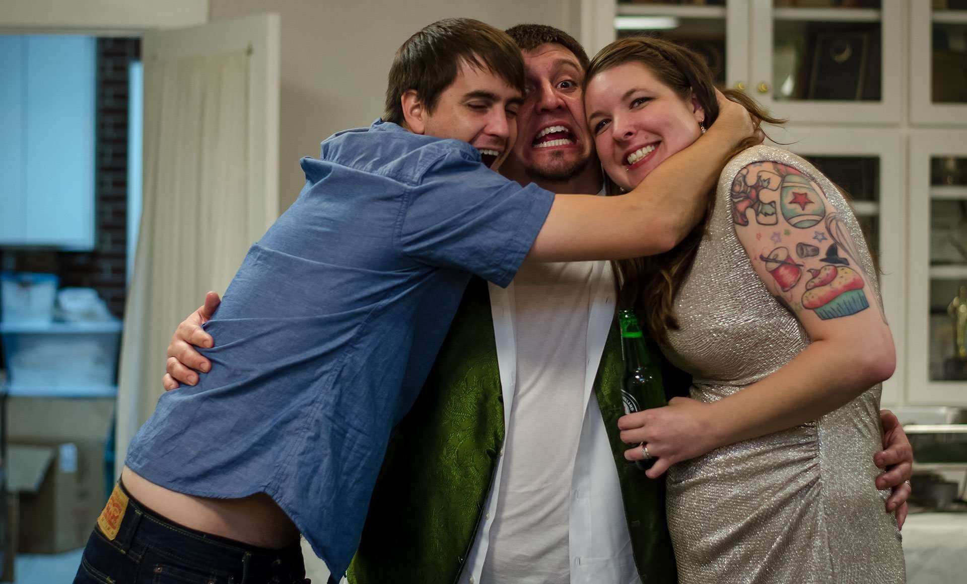 Three friends in a tight, laughing group hug indoors, woman with a colorful shoulder tattoo holding a green beer bottle