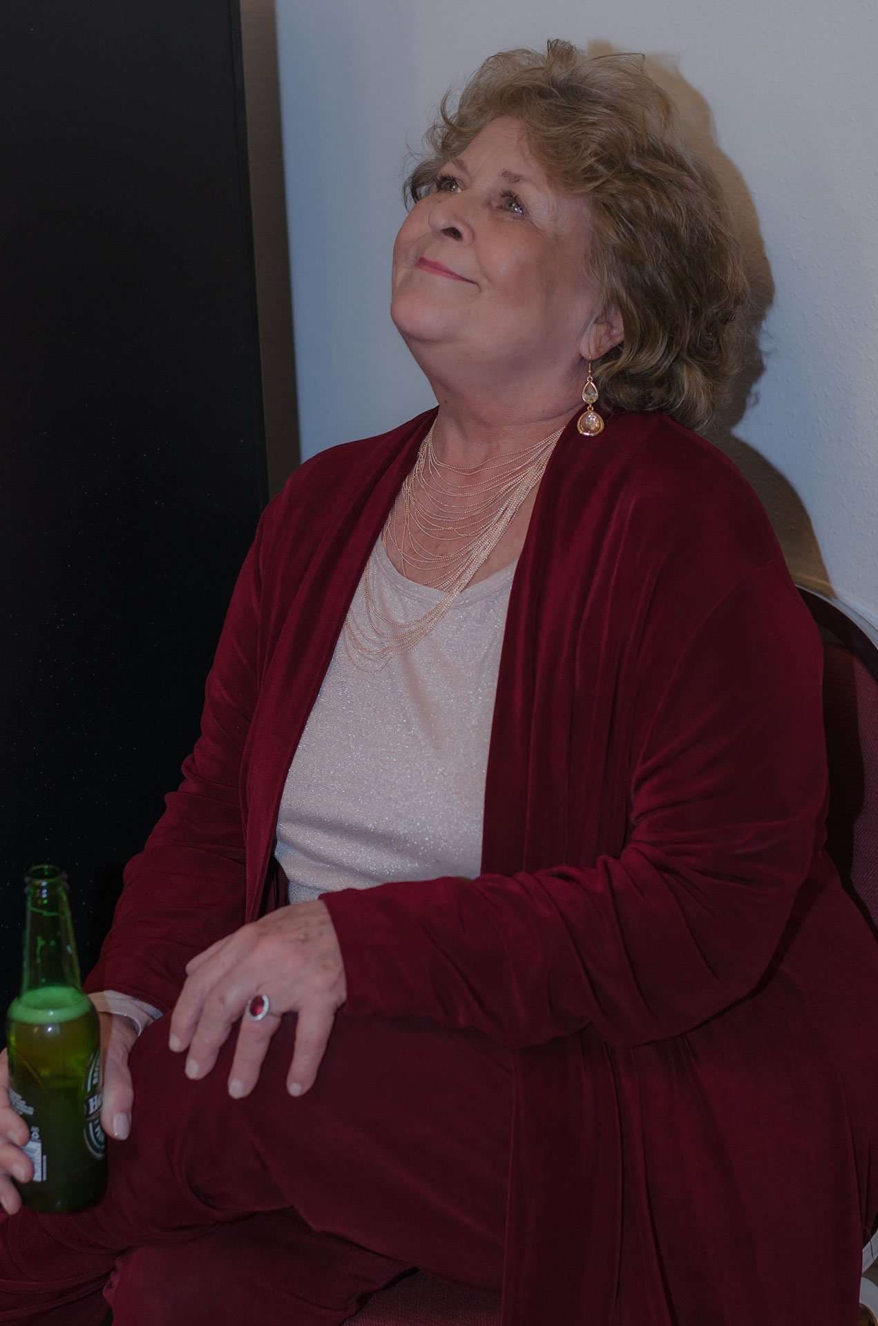 Smiling older woman in a burgundy velvet jacket and drop earrings looks upward while holding a green beer bottle