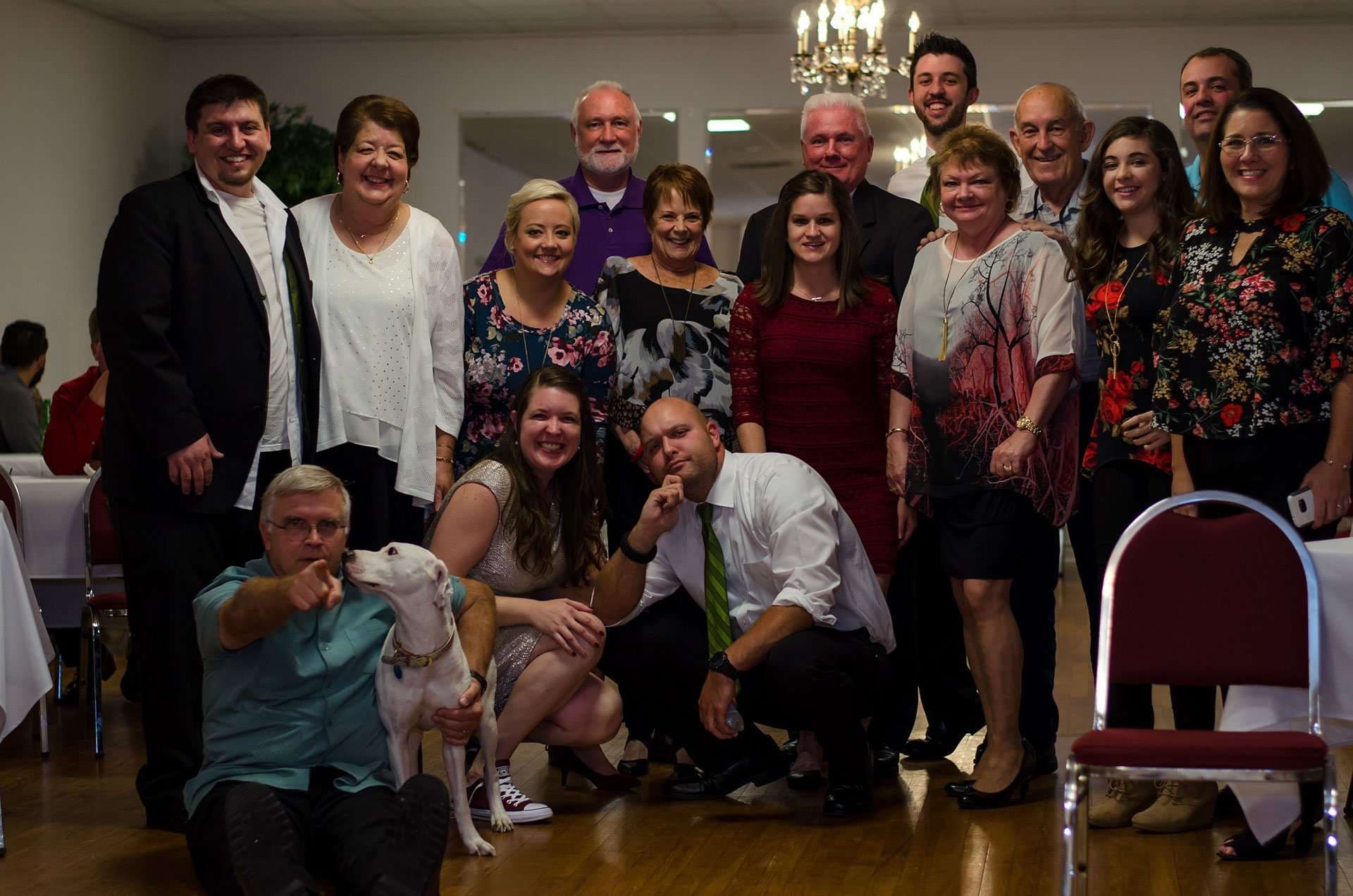 Large group of family and friends in dressy attire posing together in a hall, with a white dog in front