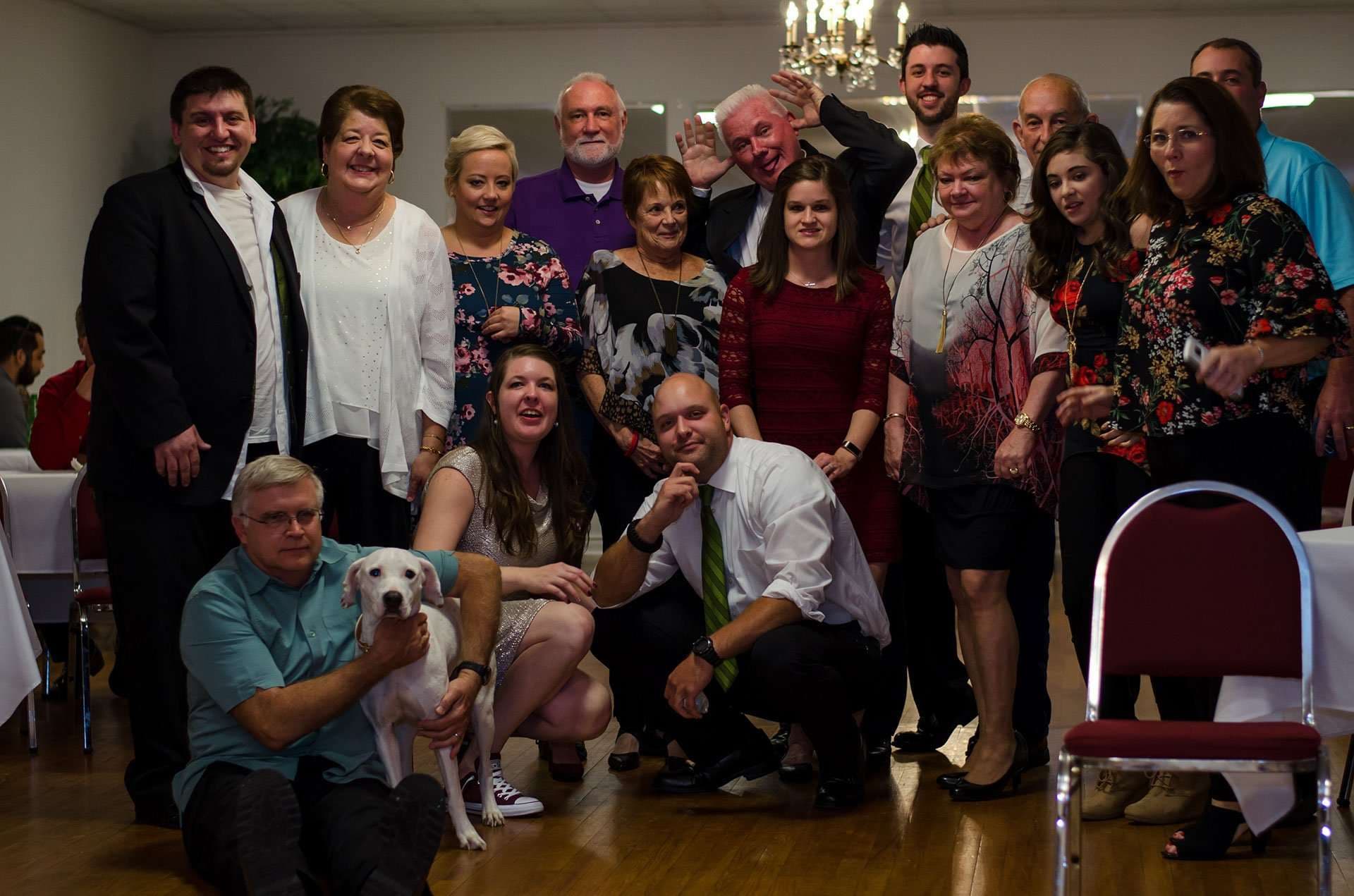 Smiling group of wedding guests posing indoors in a hall, with a white dog sitting in front beside a kneeling man
