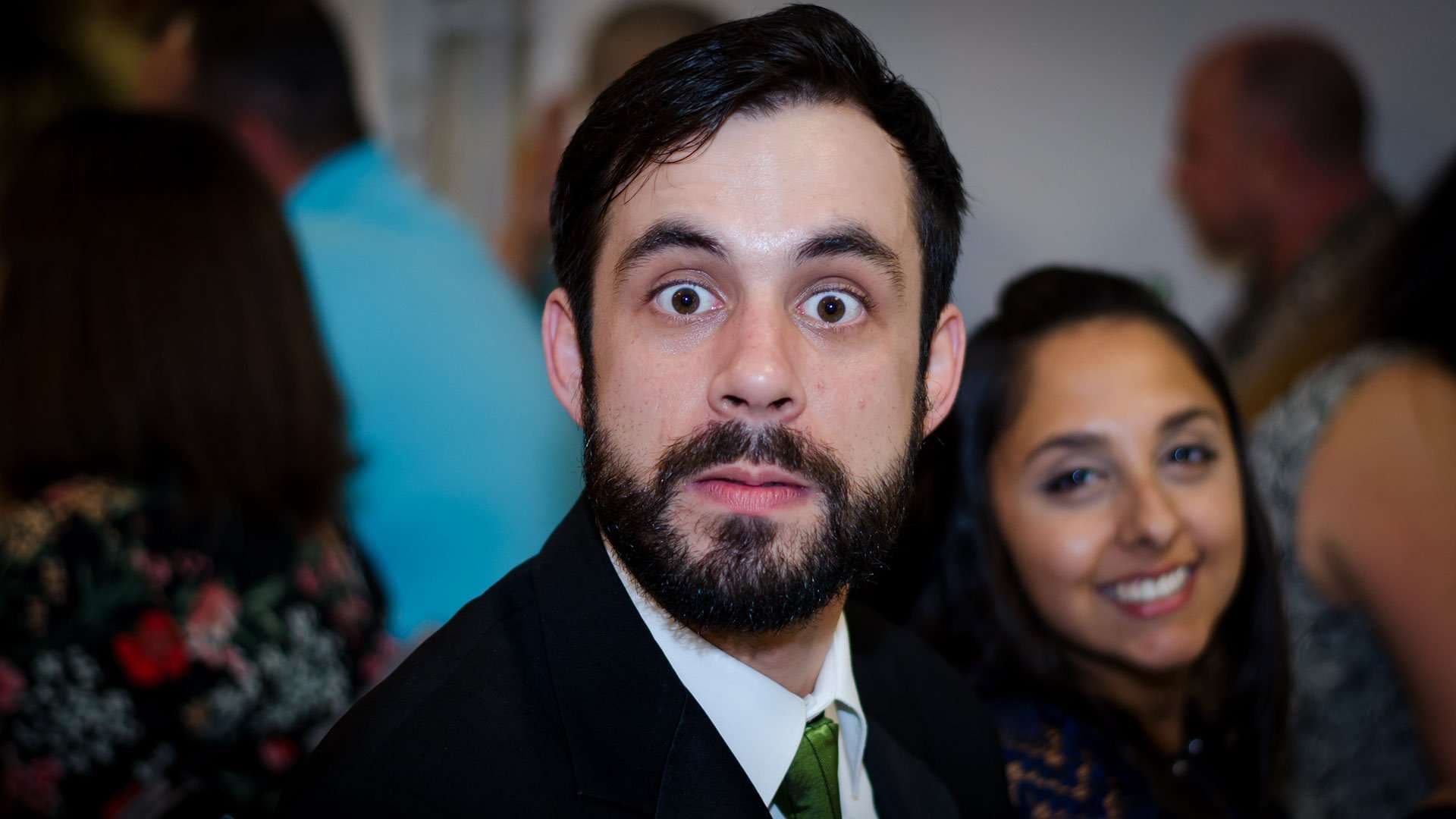 Bearded wedding guest in a suit making a wide-eyed face at the camera during the reception