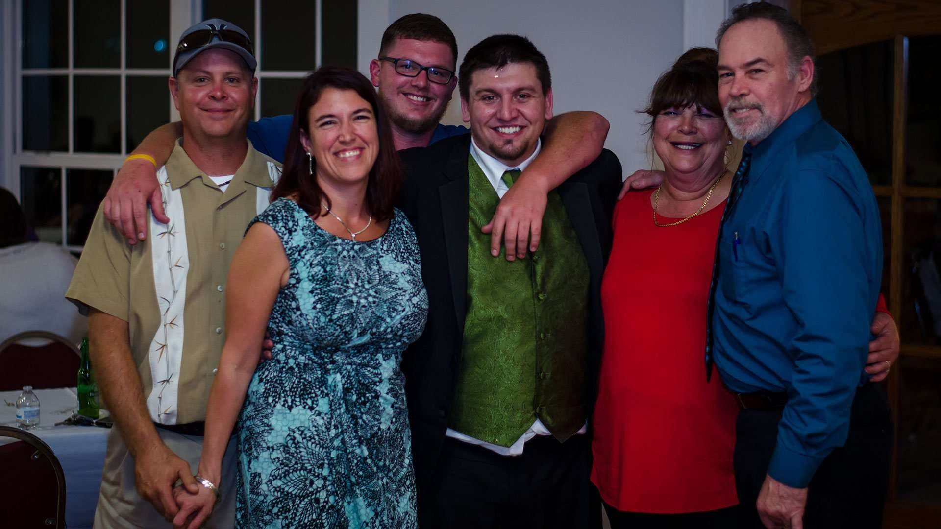 Six smiling family and friends posing arm in arm indoors, with a young man in a green vest at center