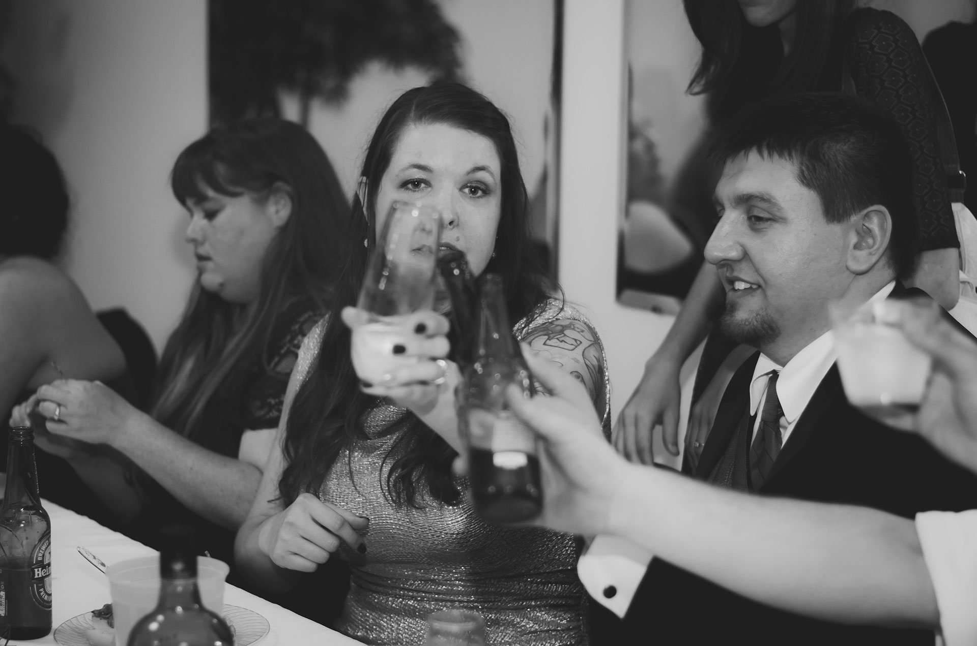 Black-and-white shot of wedding guests raising glasses and bottles in a toast at a table, a man in a suit smiling