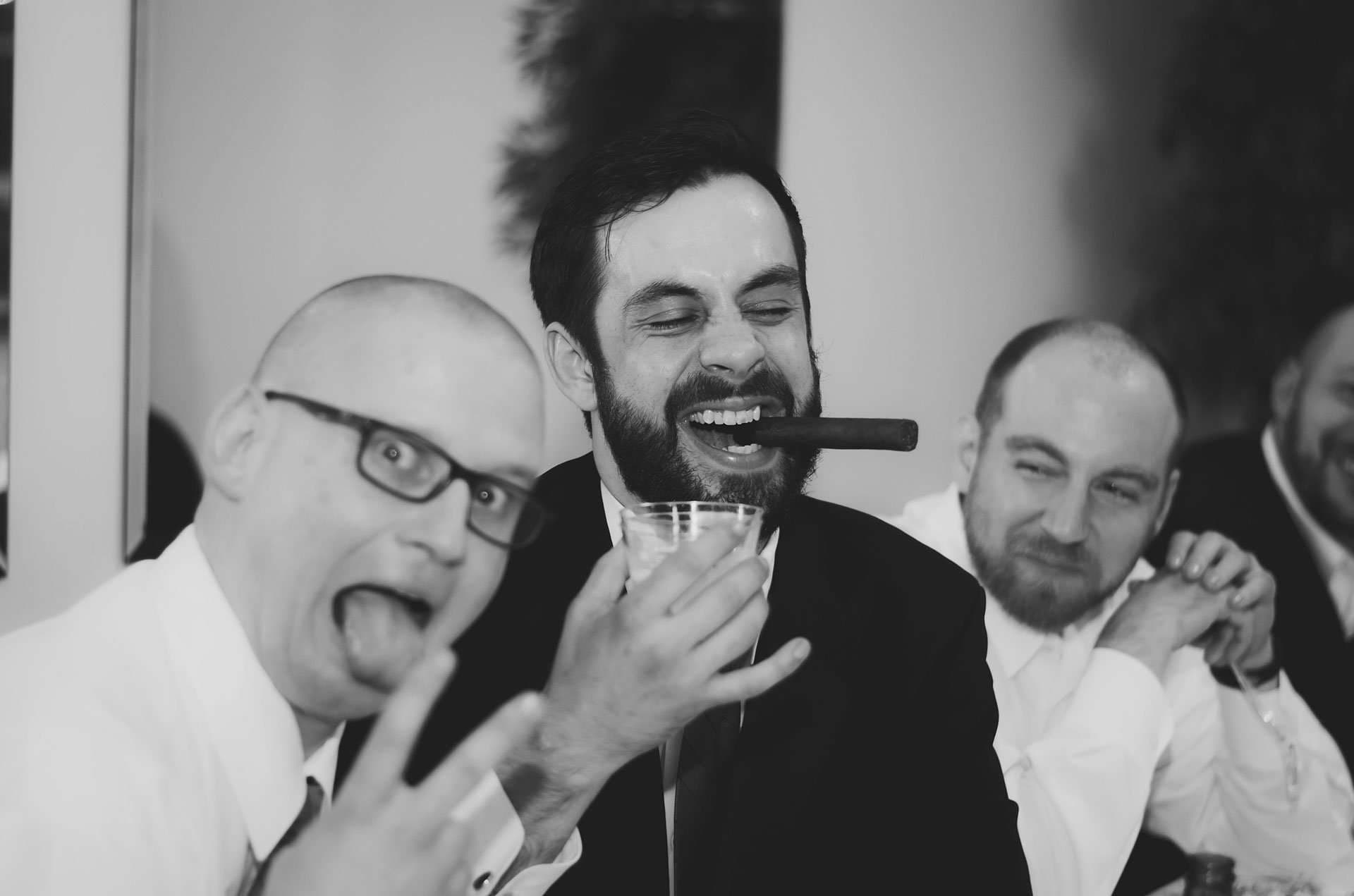 Black-and-white shot of bearded man laughing with a cigar in his mouth as a bald friend sticks out his tongue