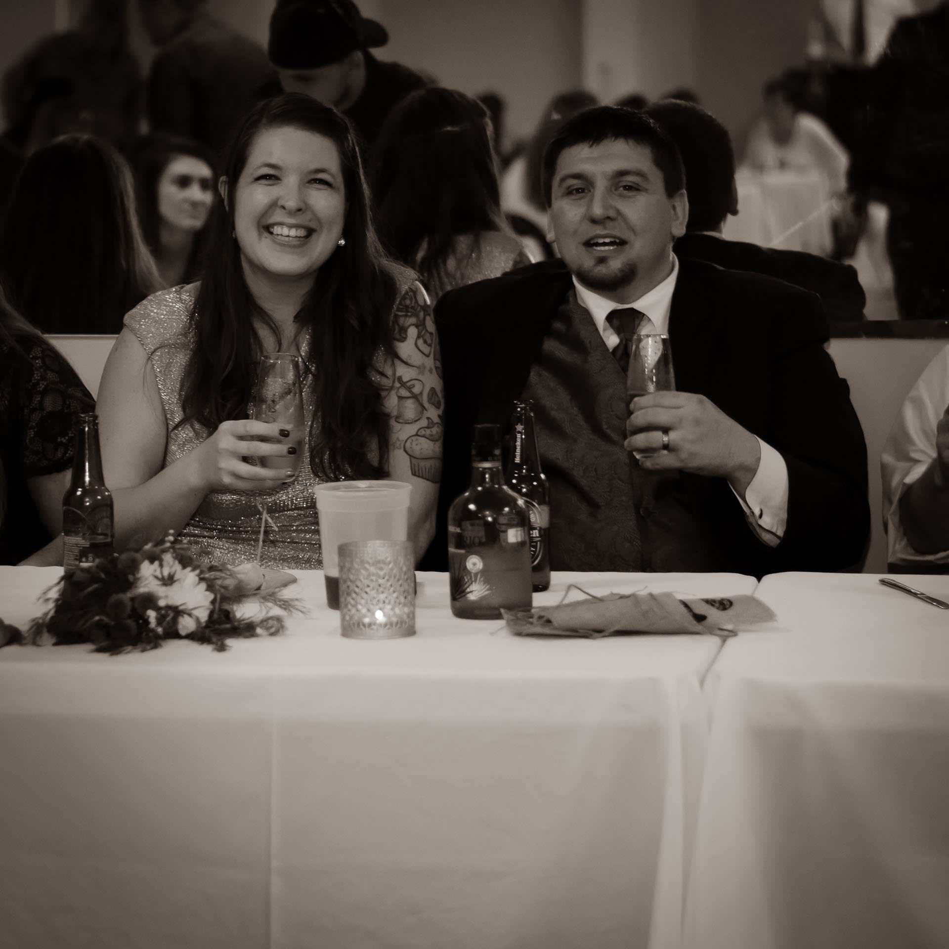 Black-and-white shot of a smiling couple seated at a reception table holding drinks, with beer bottles and guests behind